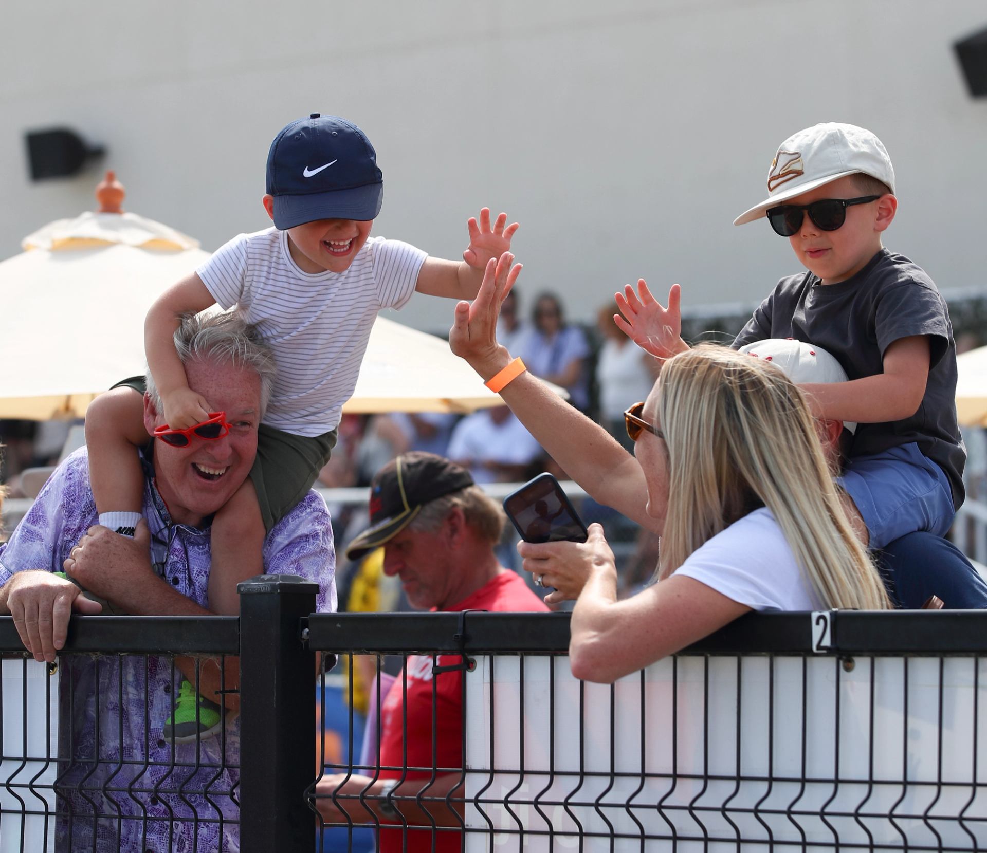 Two children reaching over a fence to high-five an adult at a racetrack event.