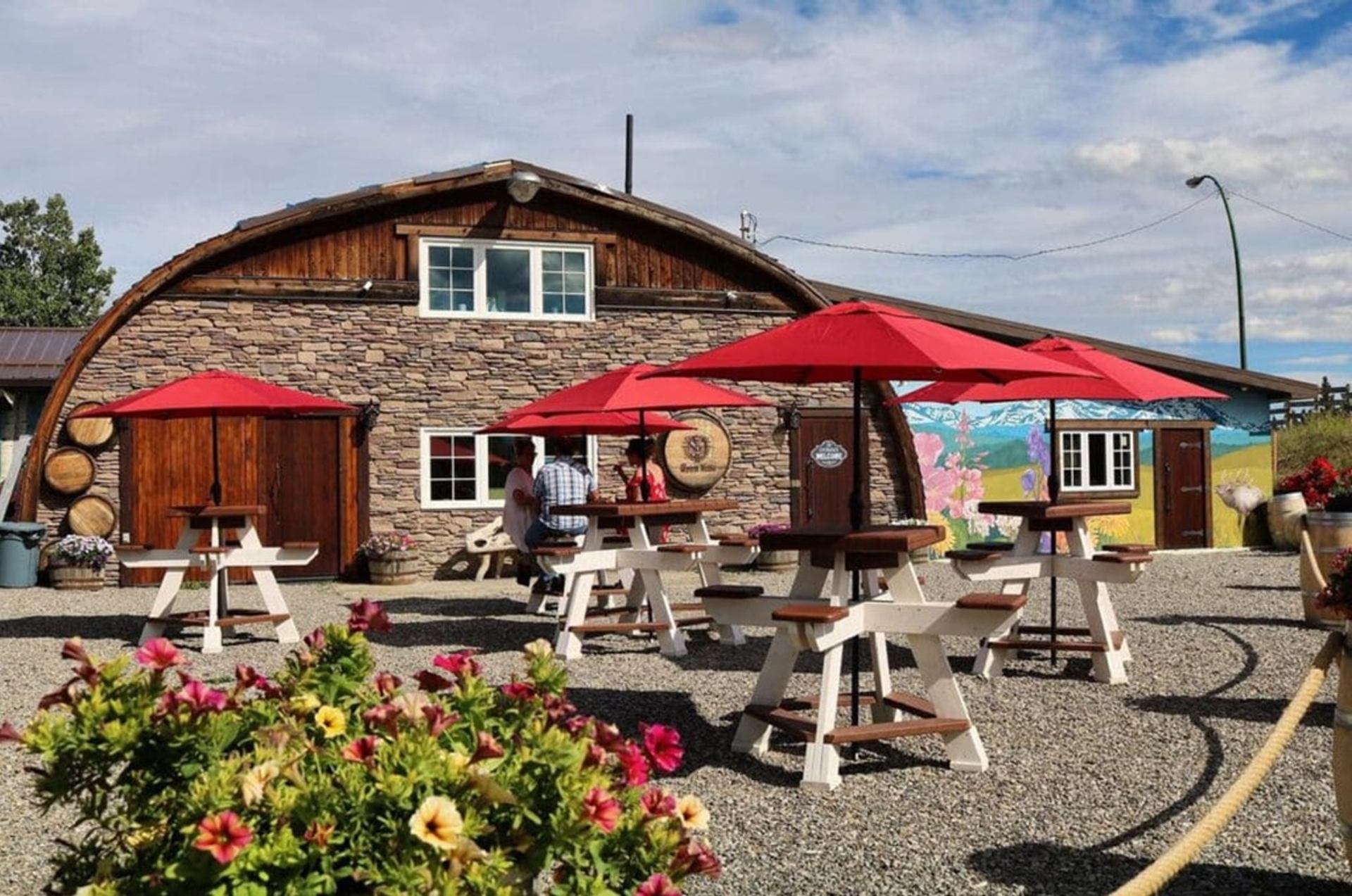 Gravel patio with picnic tables, red umbrellas and rustic stone building with mural backdrop.