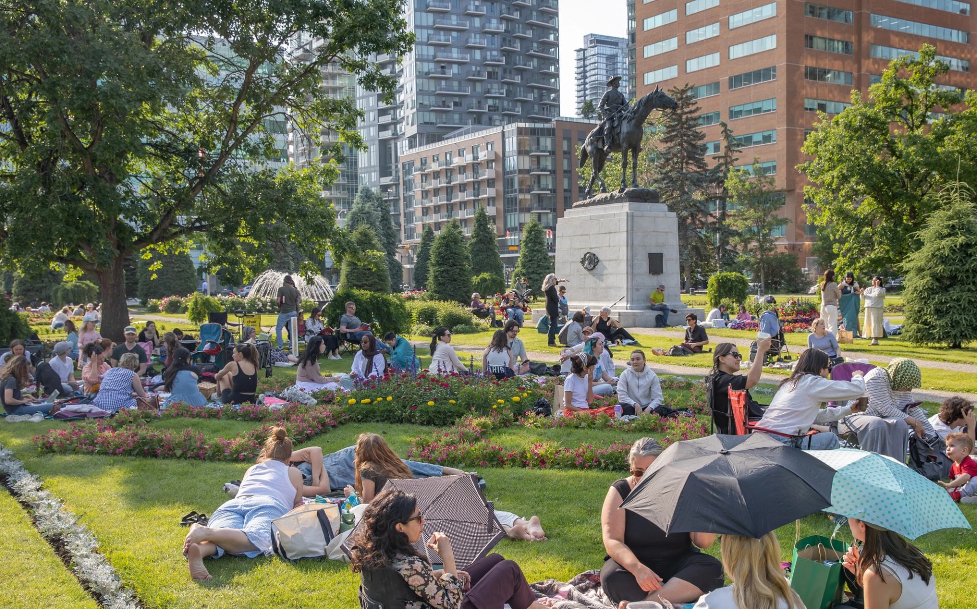 Crowds relax on park lawns near equestrian statue and city highrise buildings.