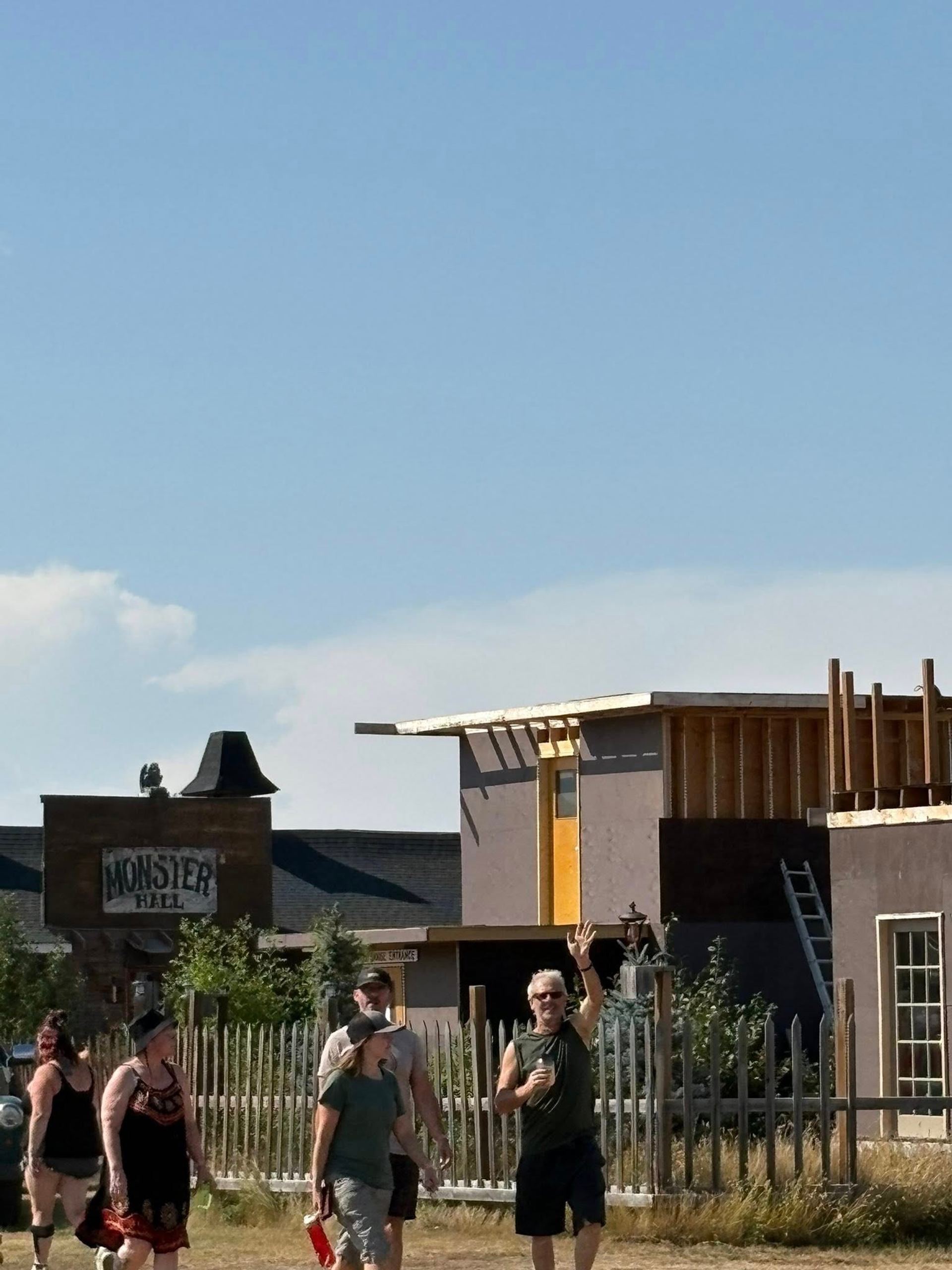 A group of people walk past a wooden fence and buildings, one labeled "Monster Hall" and another under construction, while a man waves.