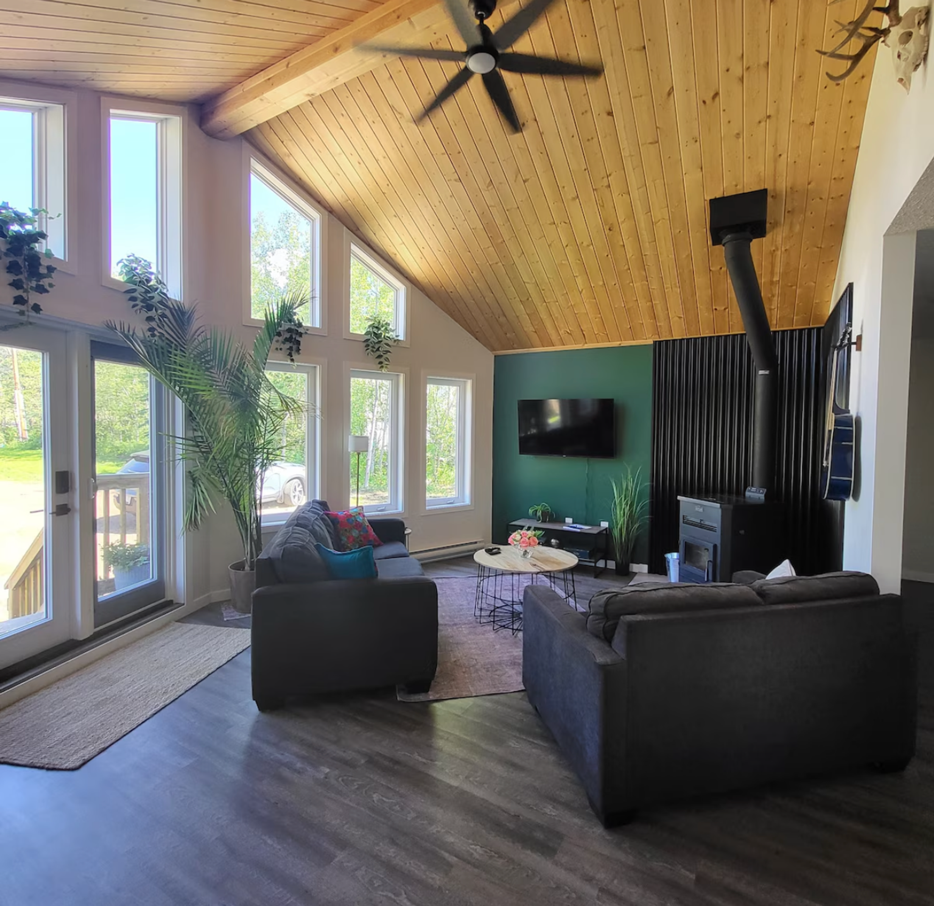 A living room with tall windows, wood ceiling, sofas, and a wood‑burning stove.