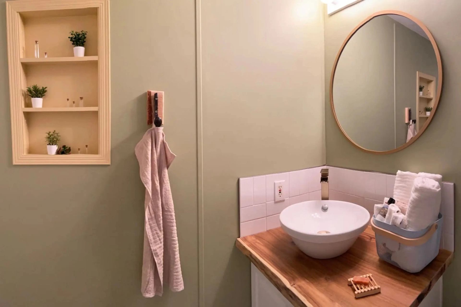 Bathroom vanity with round mirror, vessel sink, towel, shelves with plants.