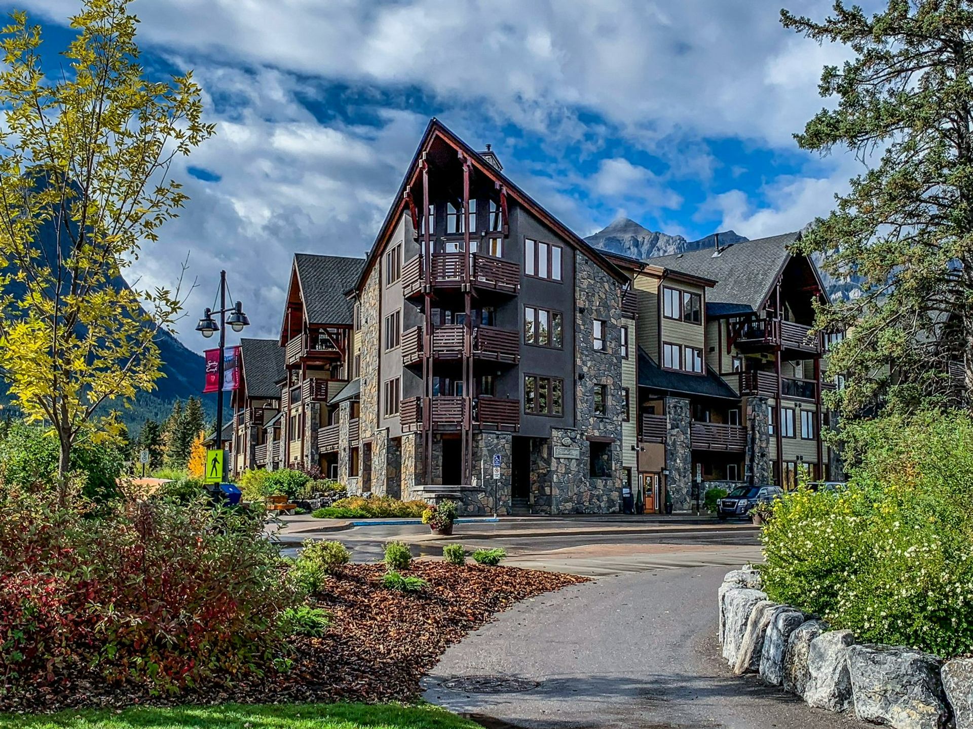 Stone and wood condominium buildings beside landscaped path, with mountains and cloudy sky.