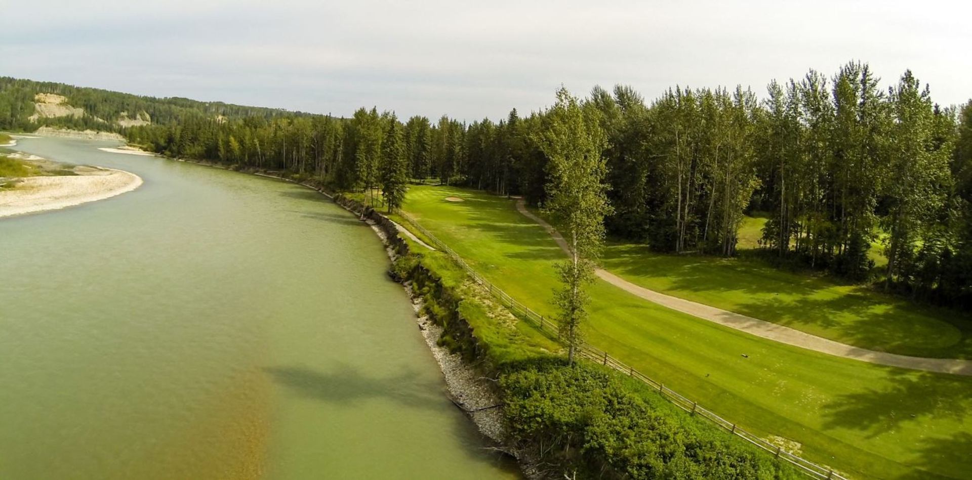 Riverside golf course with green fairways, sand traps, winding path and dense trees.