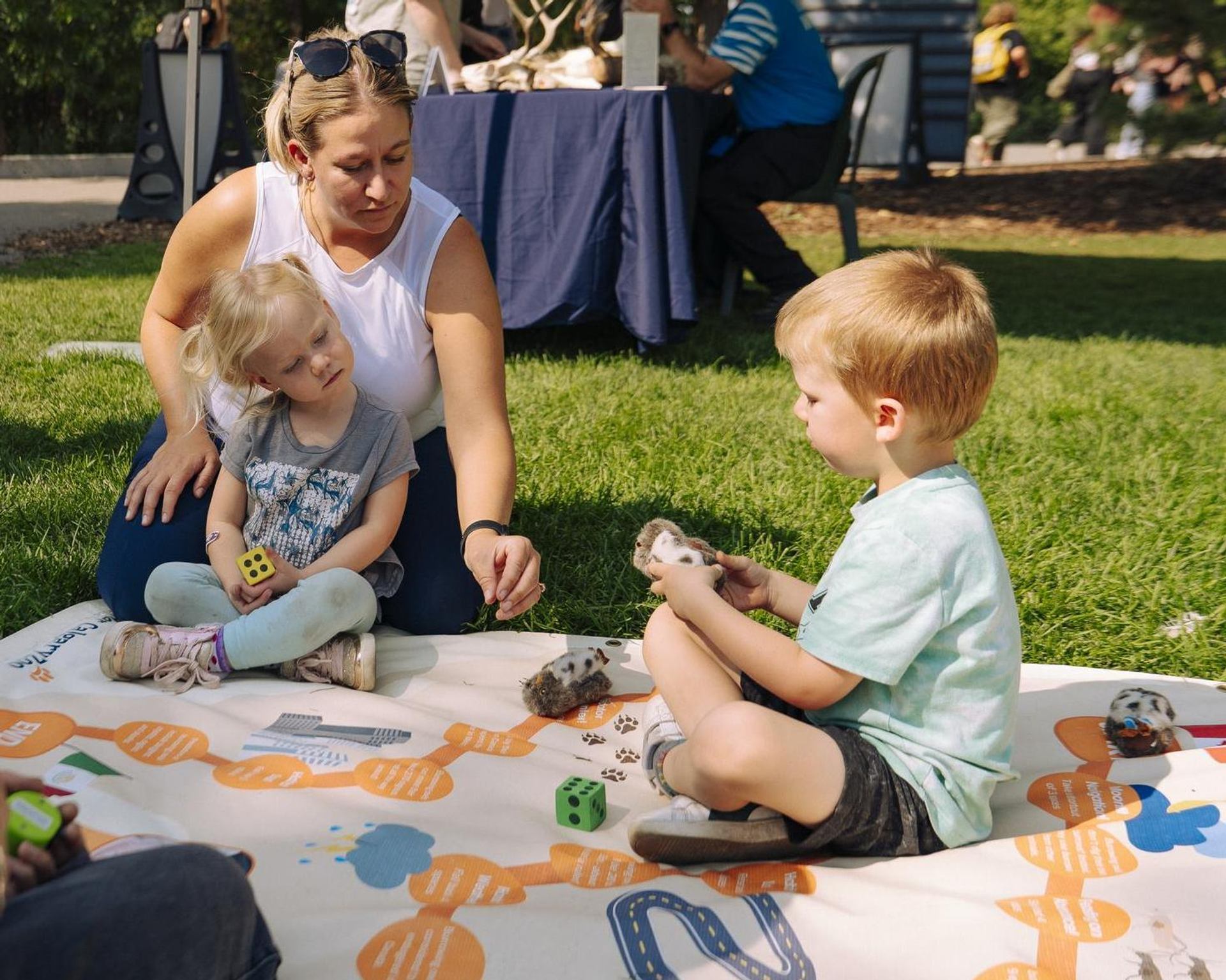An adult and two children sit on a large outdoor game mat, using dice and animal toys during a nature-themed activity.