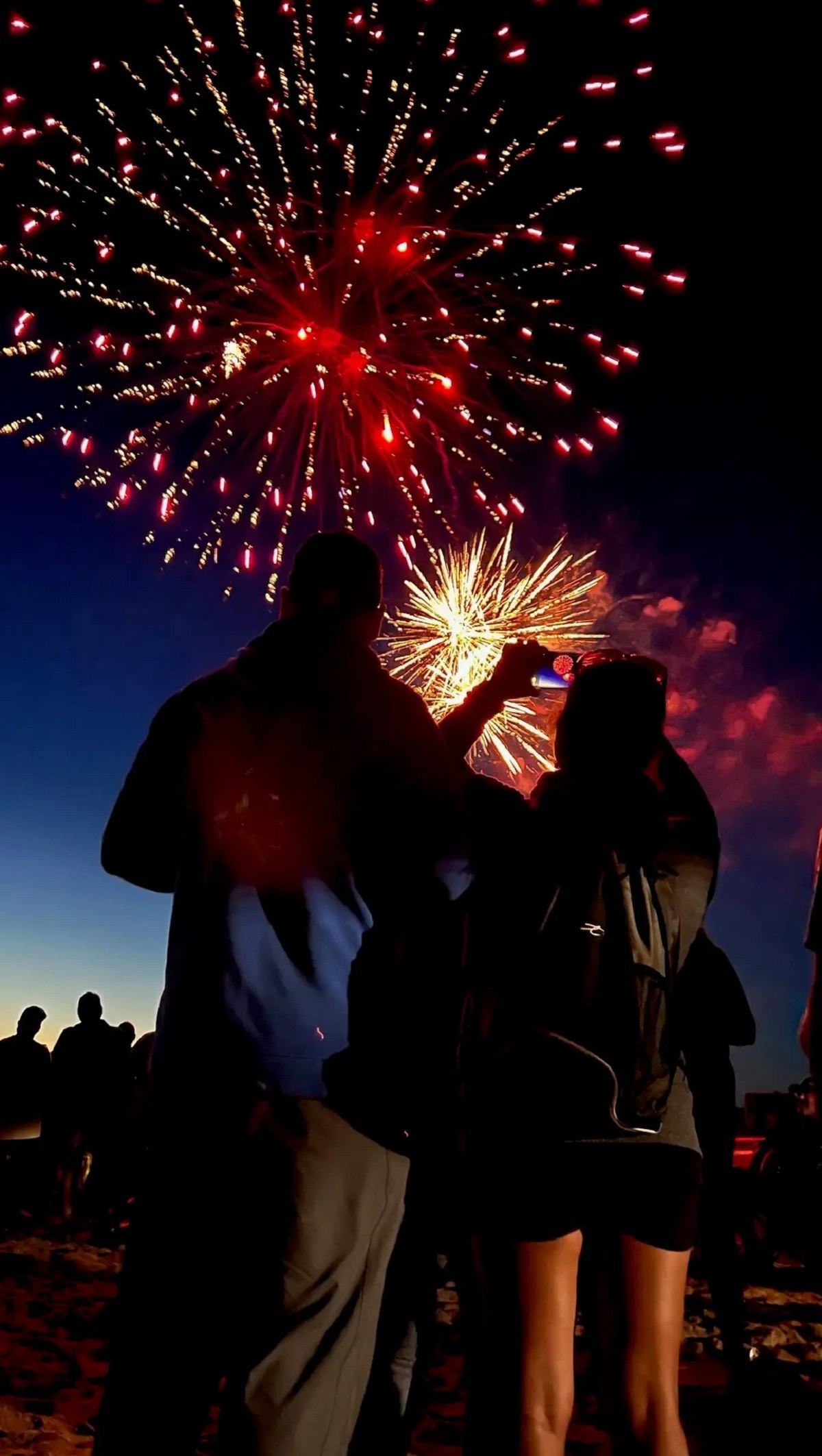 Silhouetted couple watches red and yellow fireworks light up the night sky, as a crowd gathers in celebration.