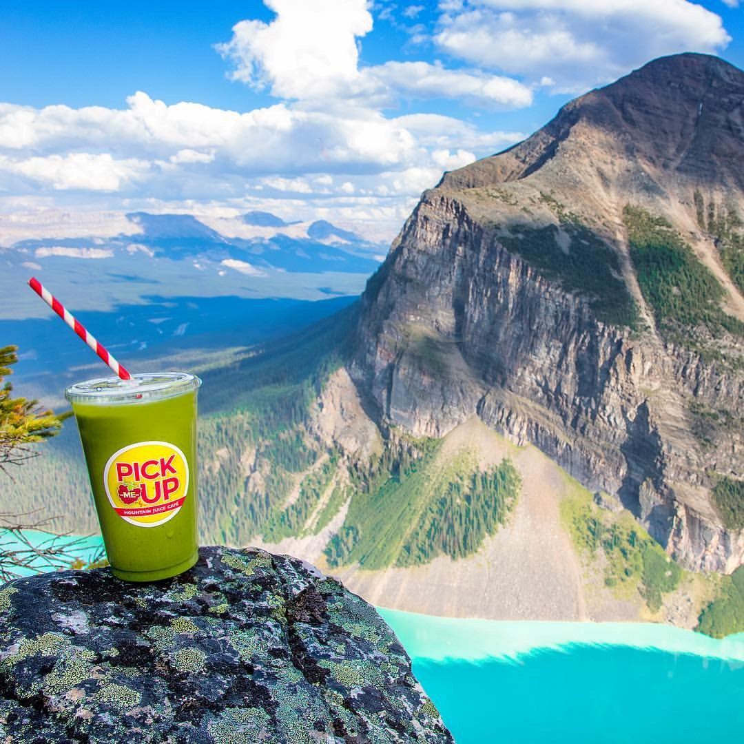 Green drink with striped straw in front of turquoise lake and rocky mountain under clear blue sky.