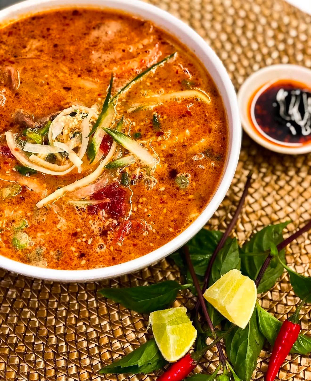 Bowl of spicy red soup with sliced vegetables, lime wedges, basil and red chilies.