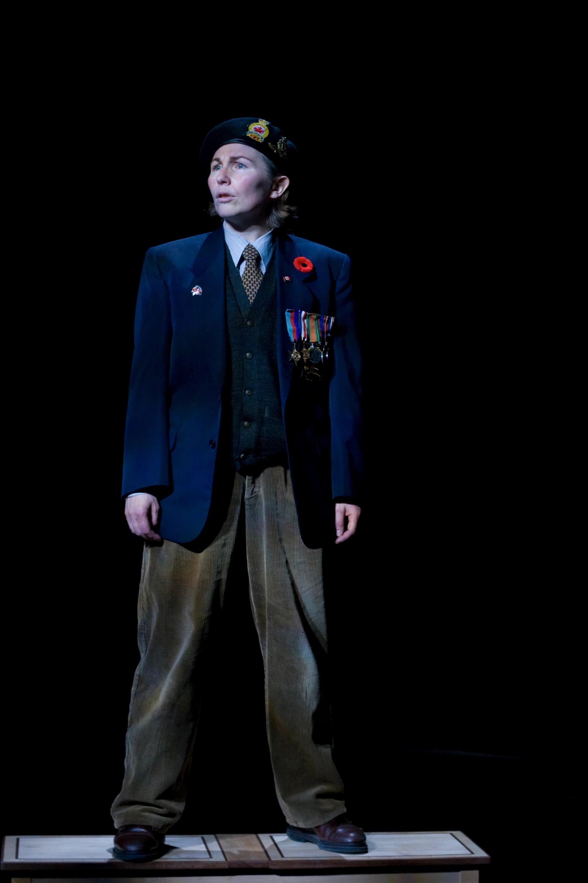 Performer in military-style blazer with medals and poppy stands onstage against black background.