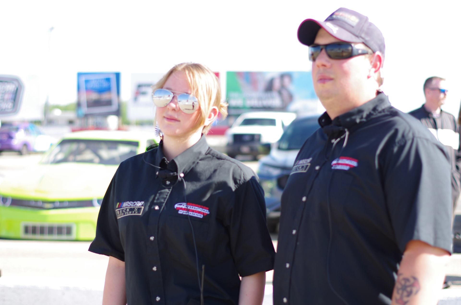 Two team members in black NASCAR shirts standing near a green stock car at the racetrack.