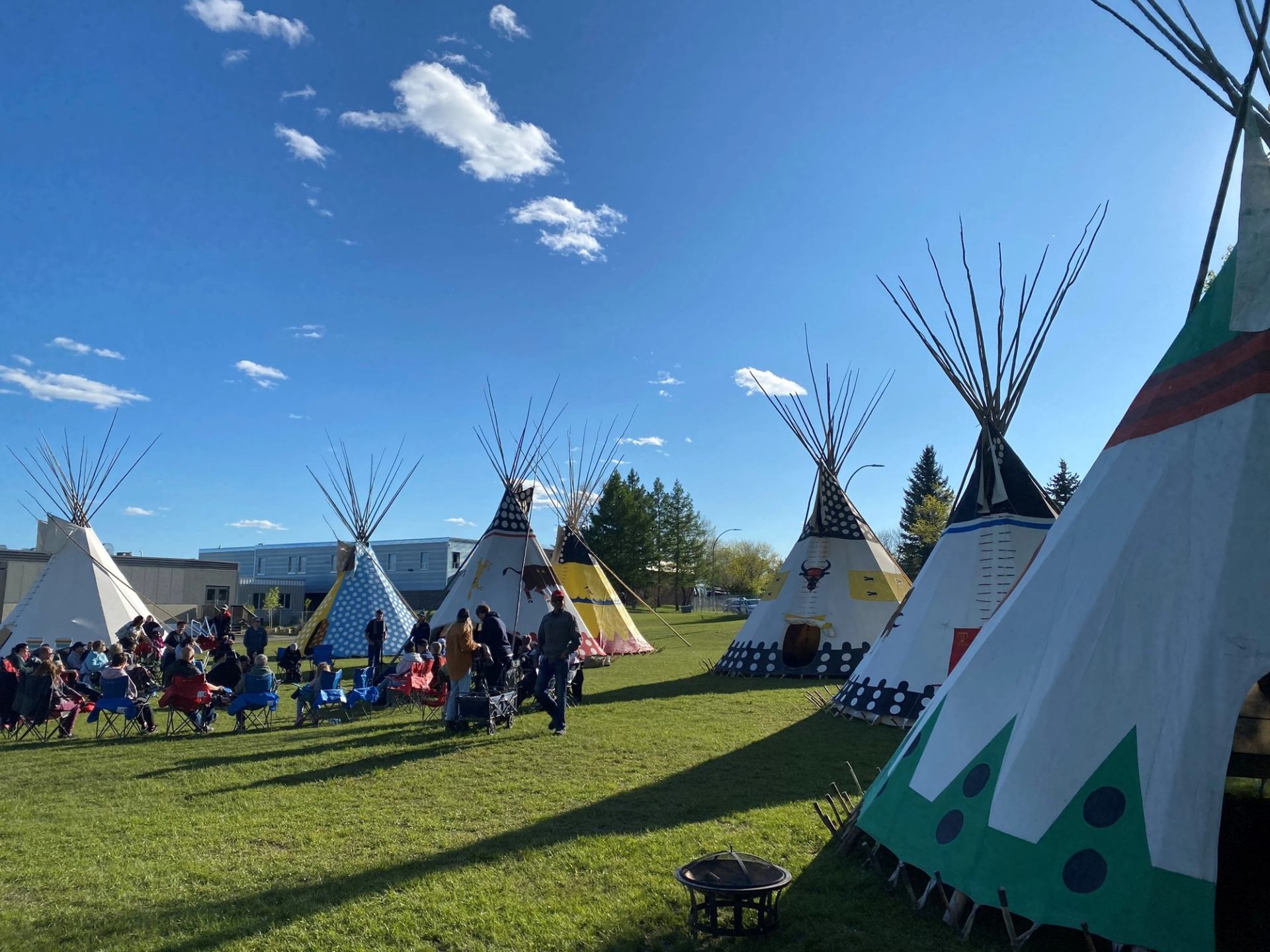 People sit on lawn chairs near painted tipis on a grassy field under blue sky.