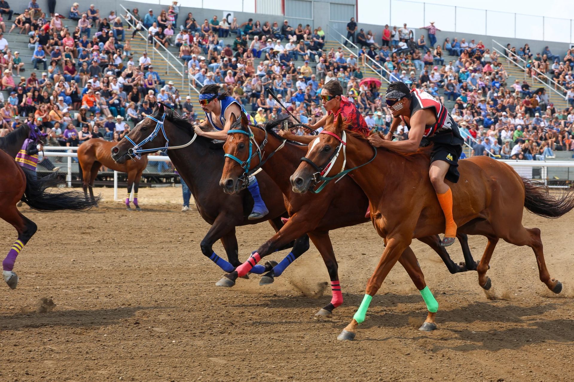 Bareback riders race horses on dirt track in front of large seated crowd.