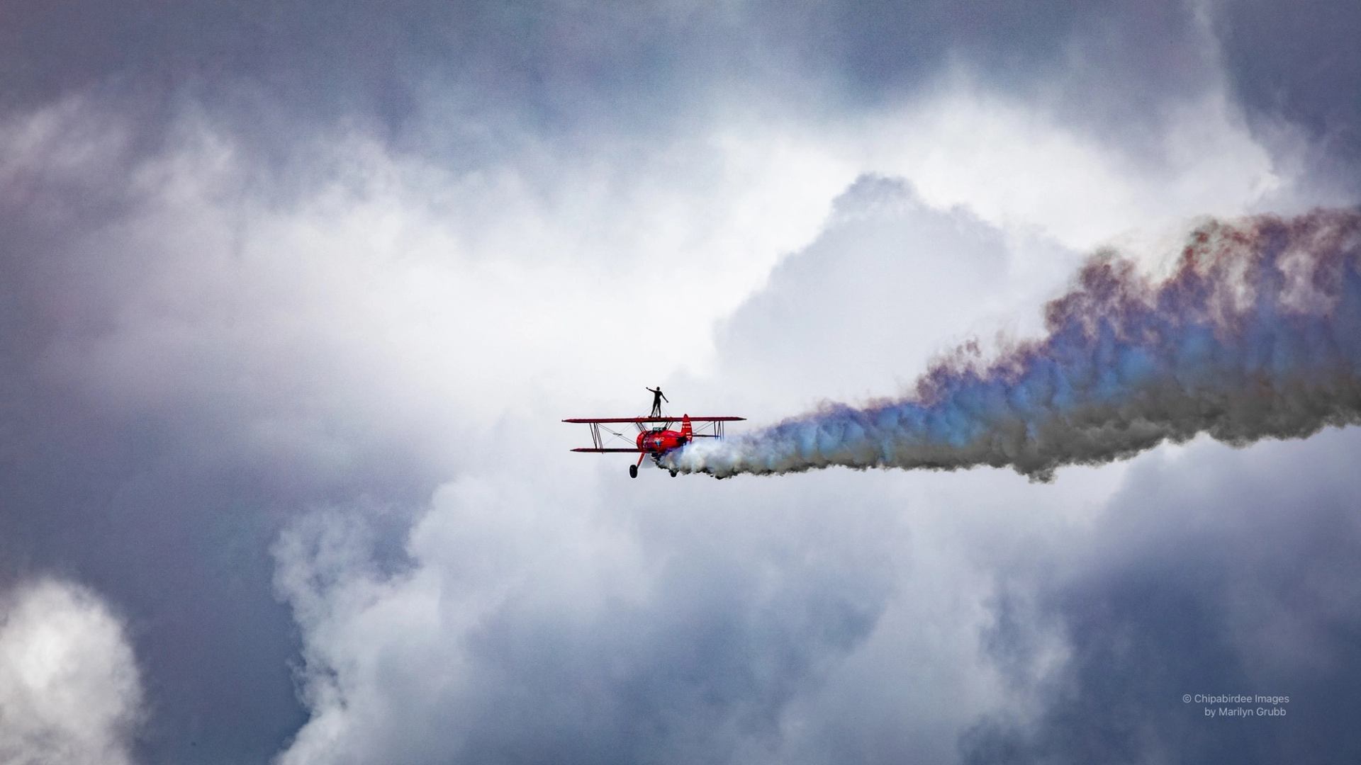 Red biplane with wing walker trailing coloured smoke against dramatic cloudy sky.