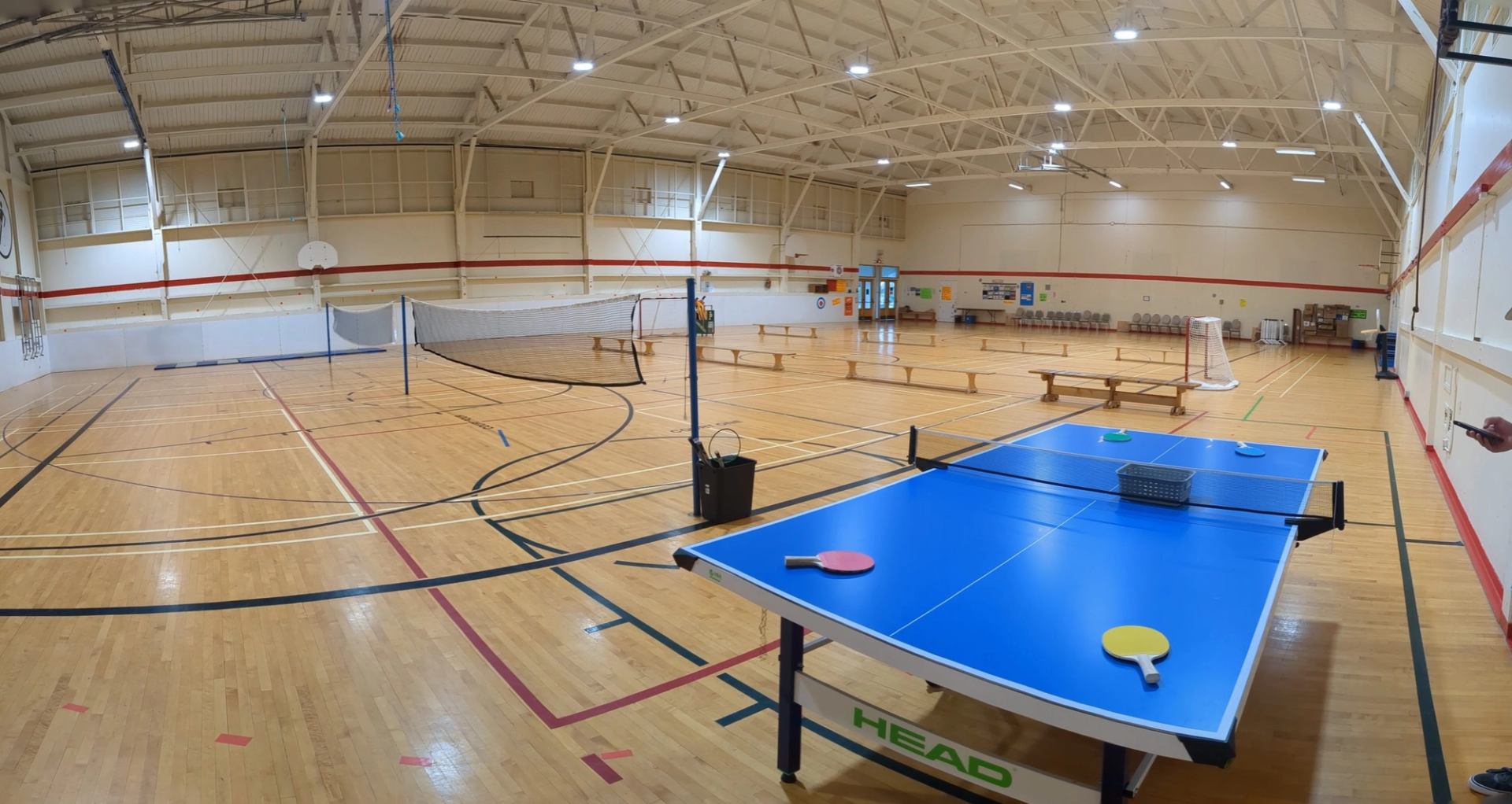 School gymnasium with table tennis setup, badminton net, benches and sports equipment on floor.