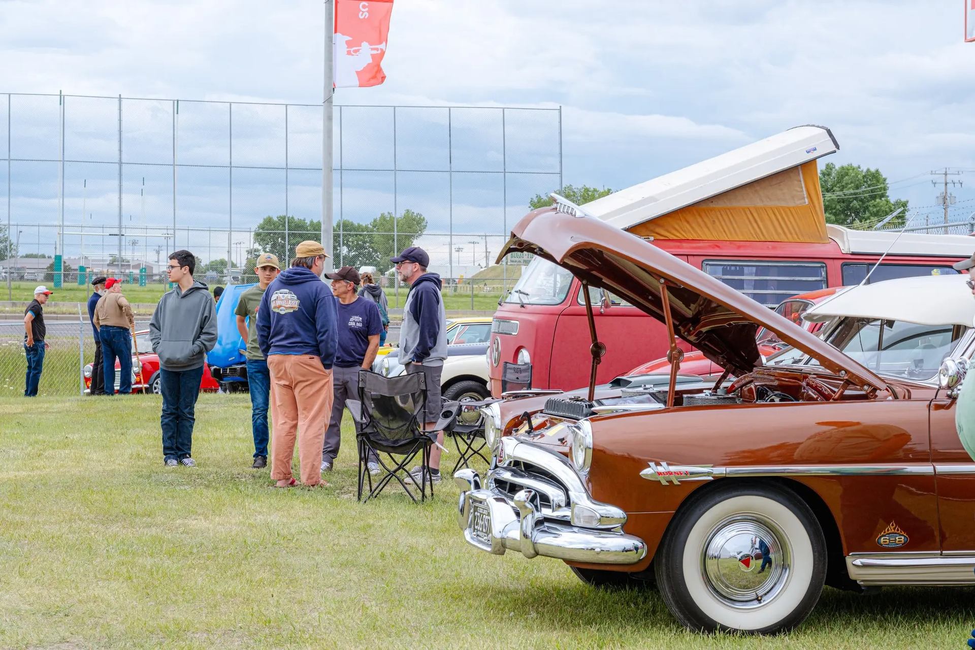Classic car with open hood at outdoor gathering beside parked vehicles and spectators near hangar fe.