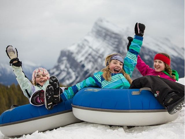 Tubing sled in snow with children posing, and snowy mountains in background at Mt Norquay.