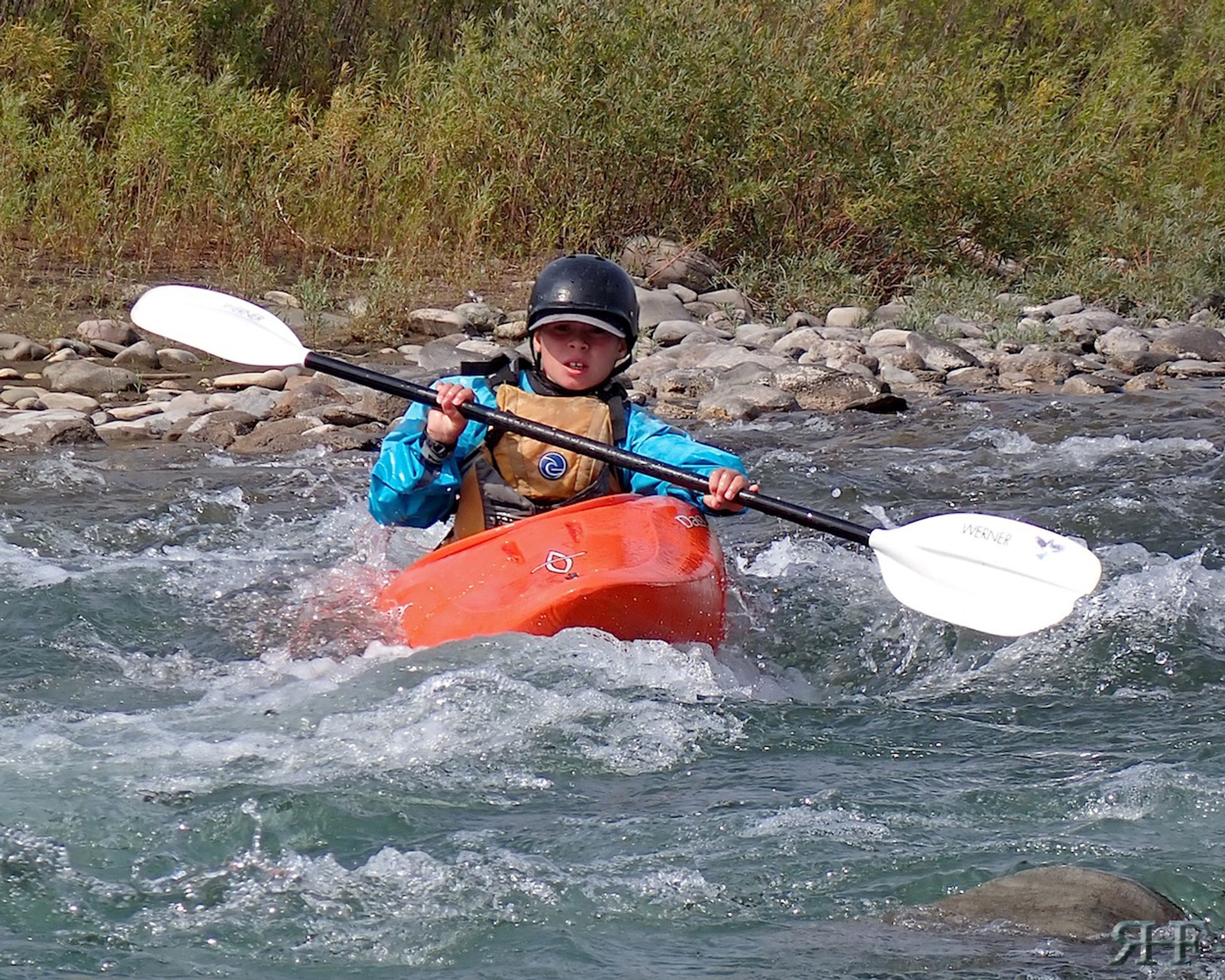 Kayaker maneuvering an orange kayak through shallow, rocky river water.