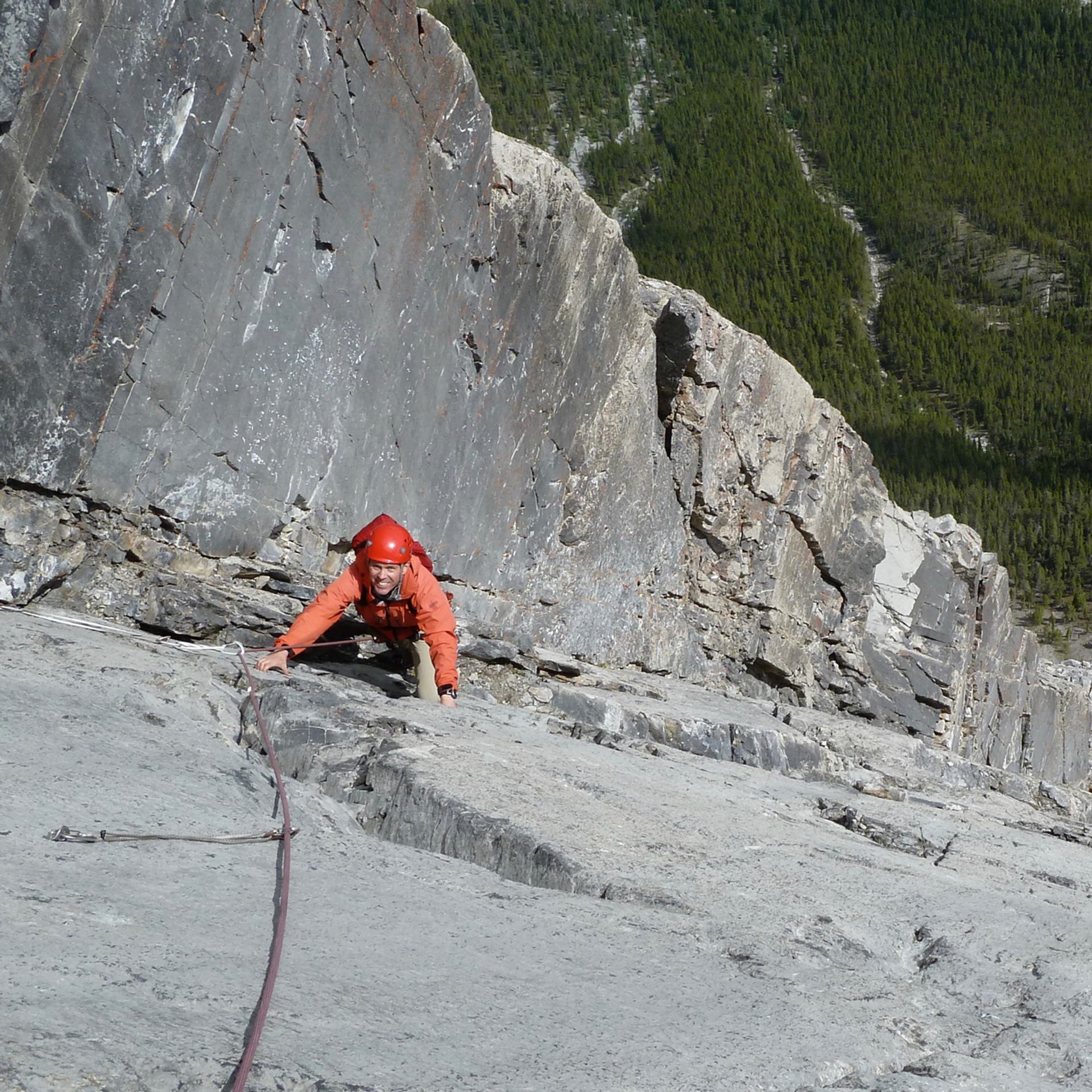 Climber in red gear scaling a steep cliff above a forested valley.