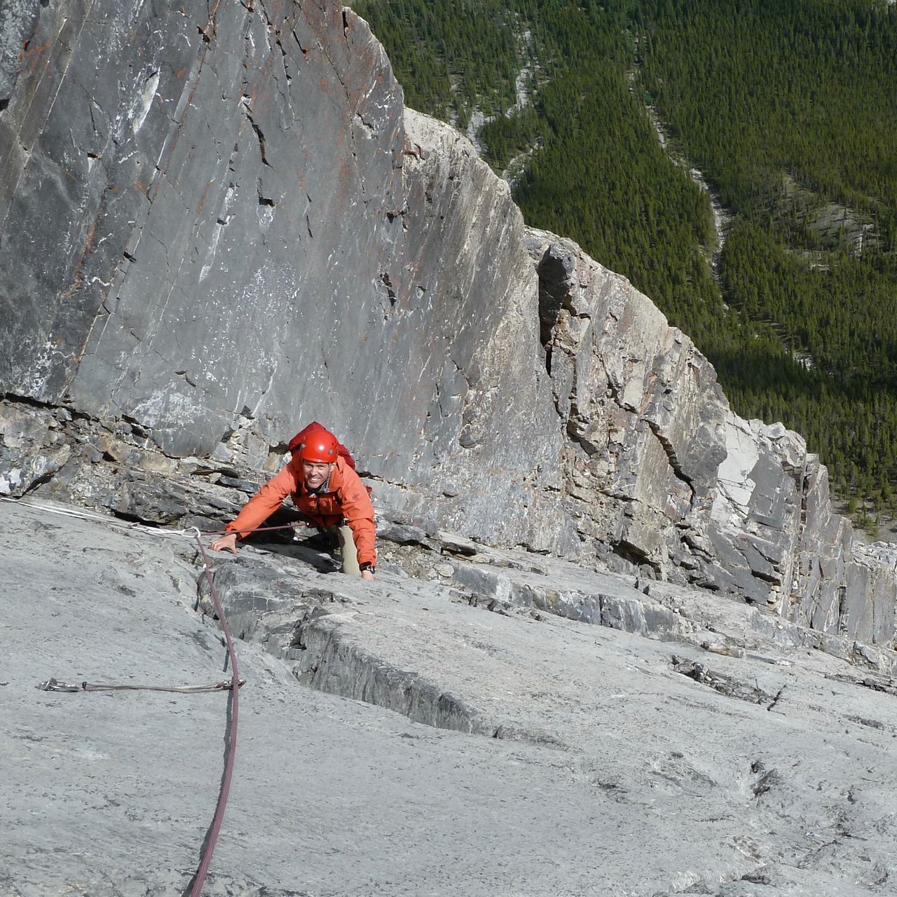 Climber in red gear scaling a steep cliff above a forested valley.