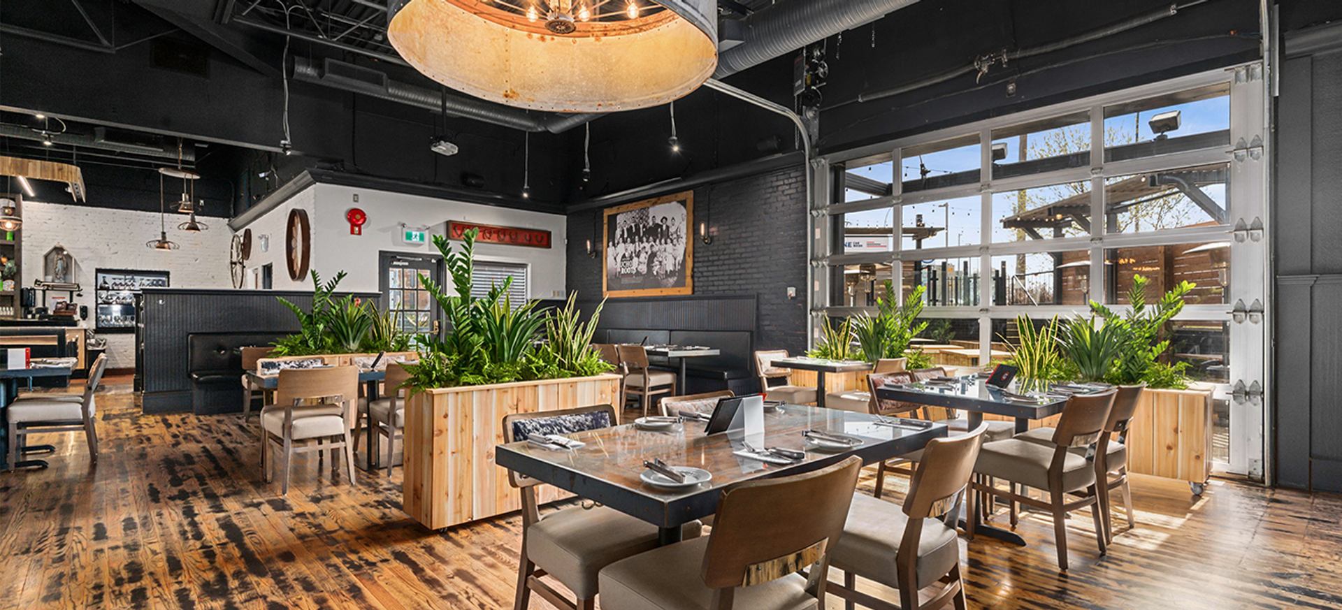 Restaurant dining room with set tables, wooden floors and large plant-filled planters.
