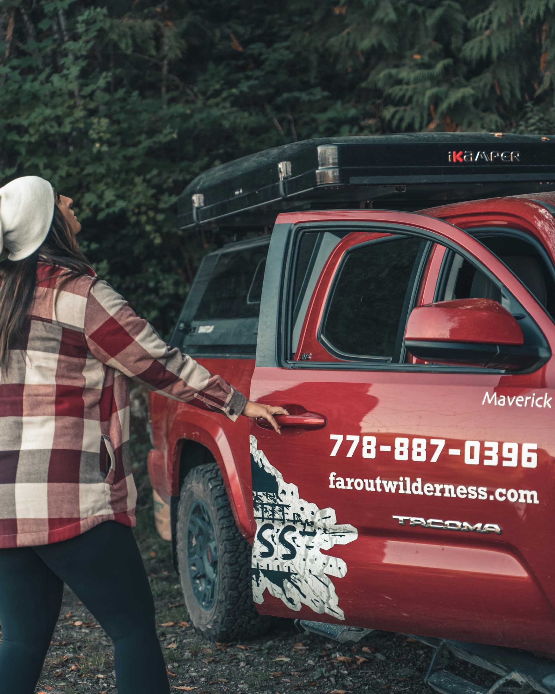 Person standing by a red truck with FallOut Wilderness branding and roof gear in a forest setting.