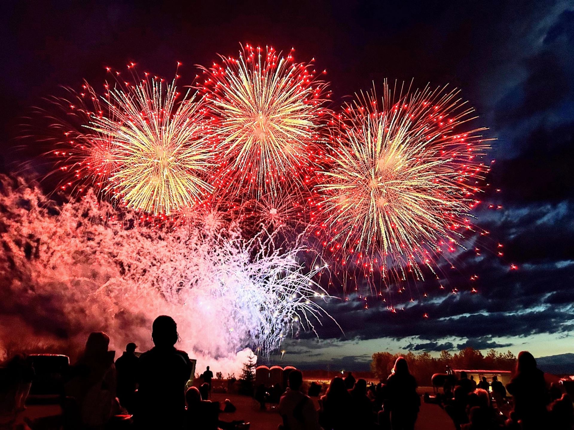 Crowd watches large red and white fireworks bursting over a dark evening sky.