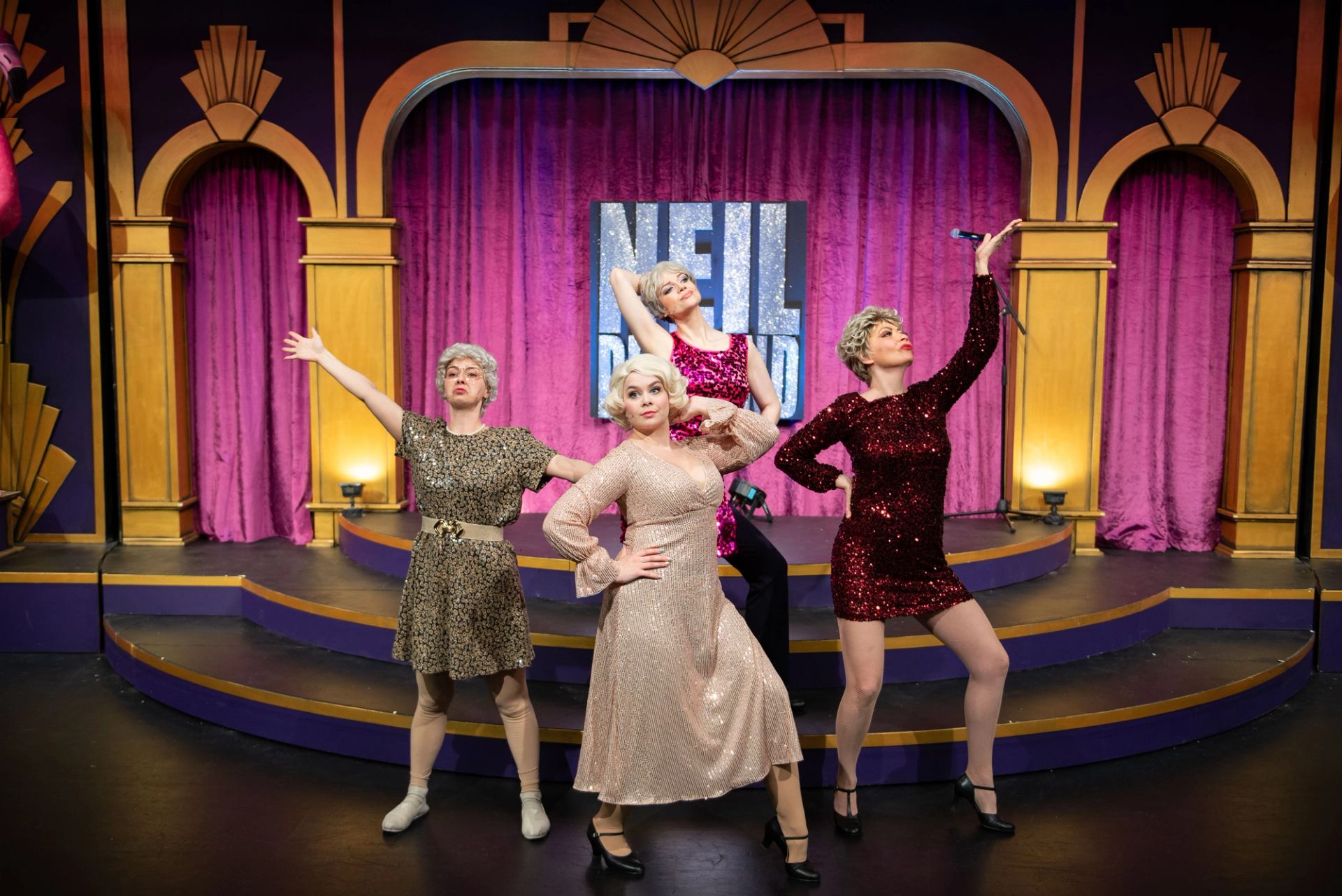 Four performers in sequined dresses pose dramatically on a theatre stage with purple curtains.