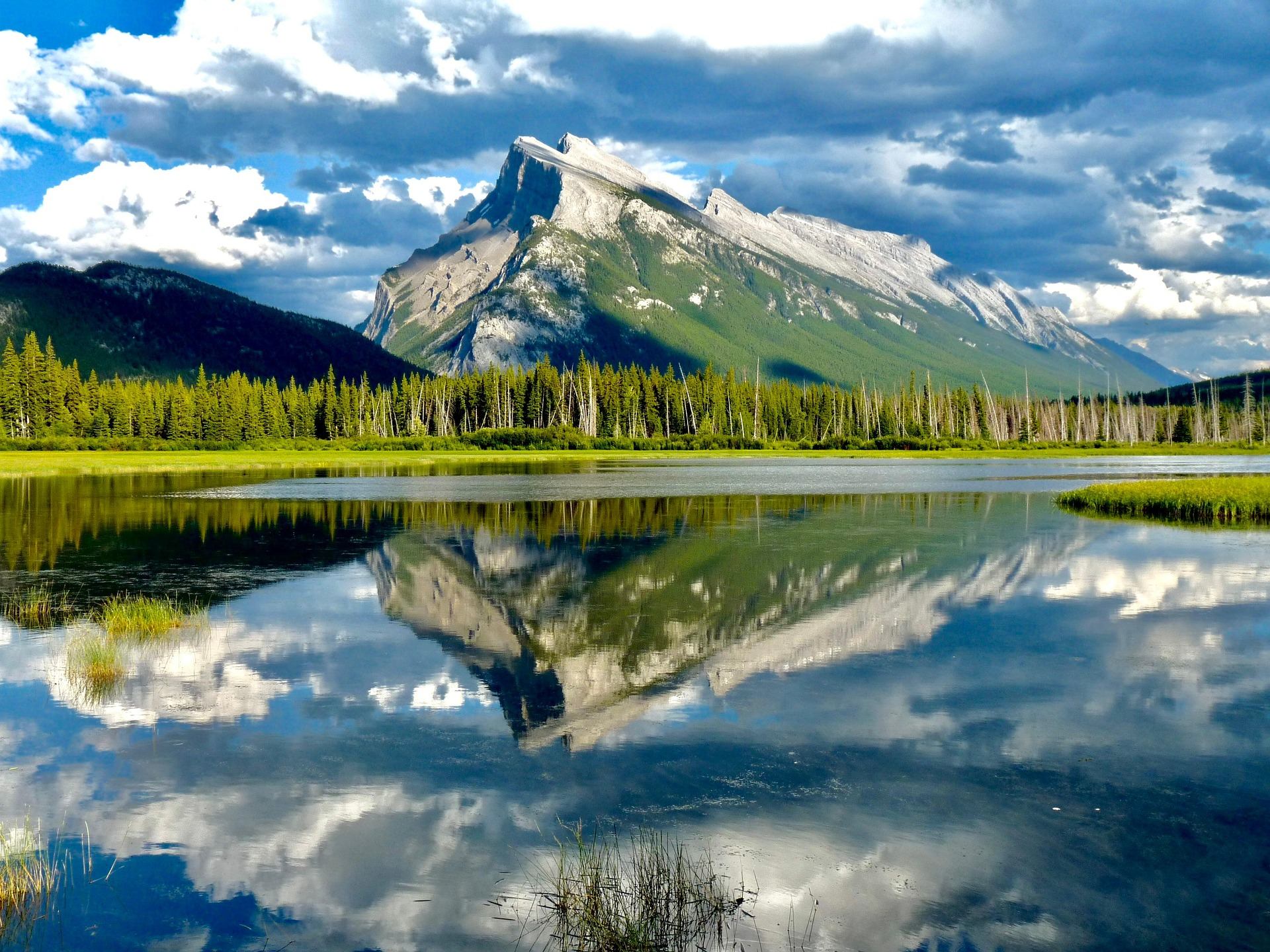 Mountain range and forest reflected in calm lake under partly cloudy blue sky.