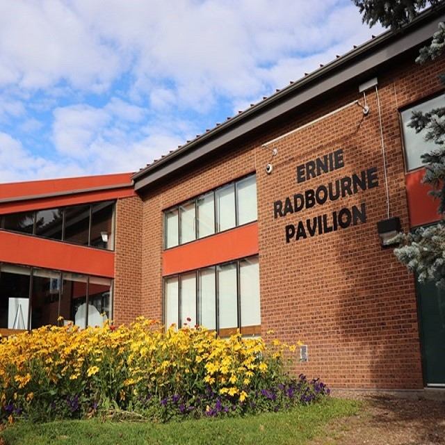 Brick building with windows and sign reading “Ernie Raddbourne Pavilion,” with flowerbeds in front.
