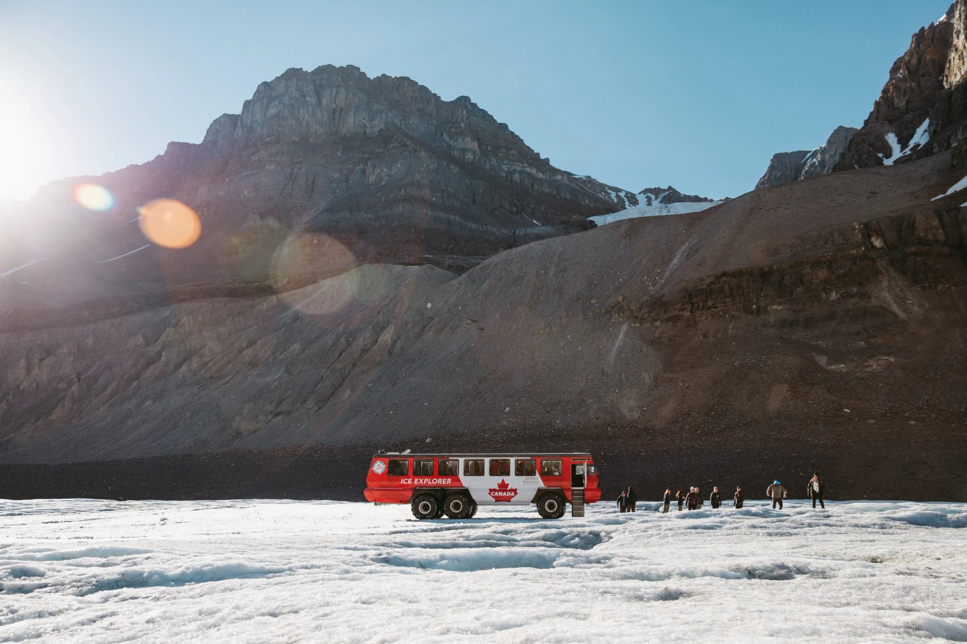 Red Ice Explorer bus and visitors stand on snowy glacier below rocky mountain slope.