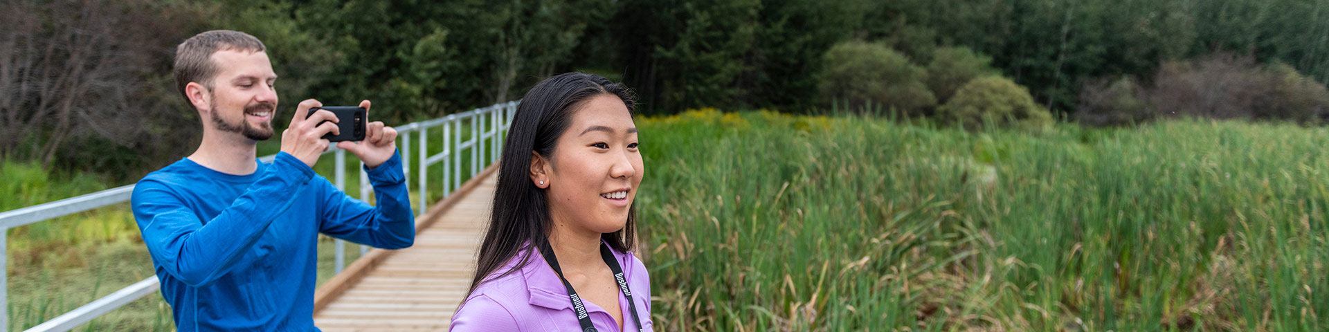 Two visitors stand on a boardwalk beside wetland vegetation, with one taking a photograph.