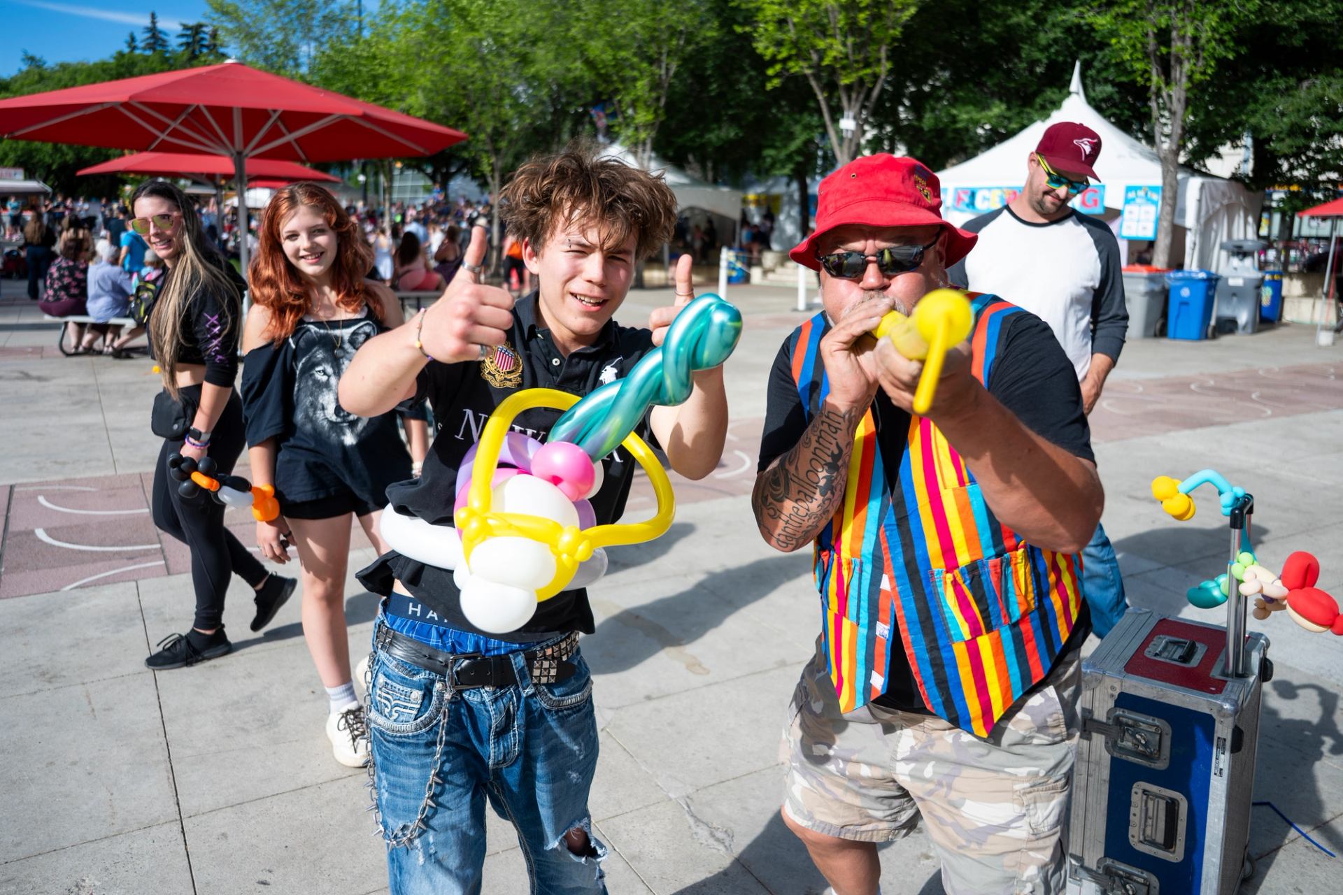 Street performer makes balloon animals as people with balloon bracelets smile in crowd.