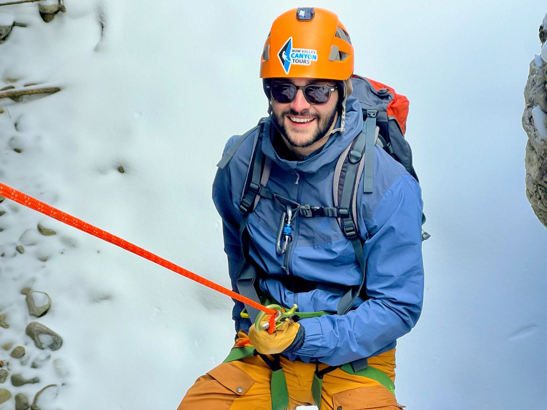 Adventurer in helmet and gear rappelling on snowy terrain with bright orange rope.