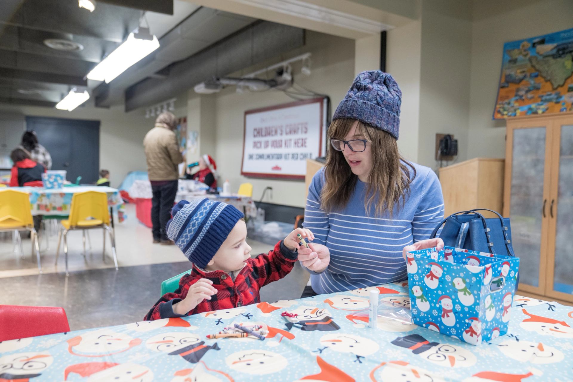 Child and adult make crafts at a holiday-themed table covered with snowman print.