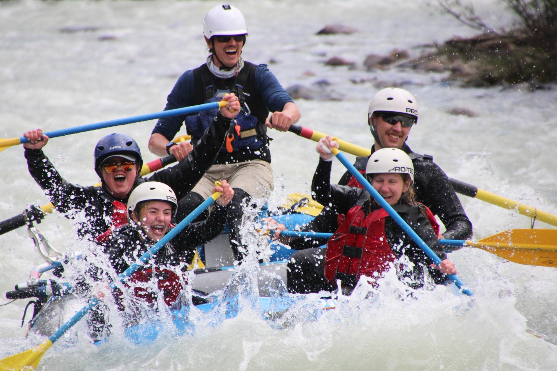 Group of rafters wearing helmets paddle through churning white water, cheering as waves crash over the raft.