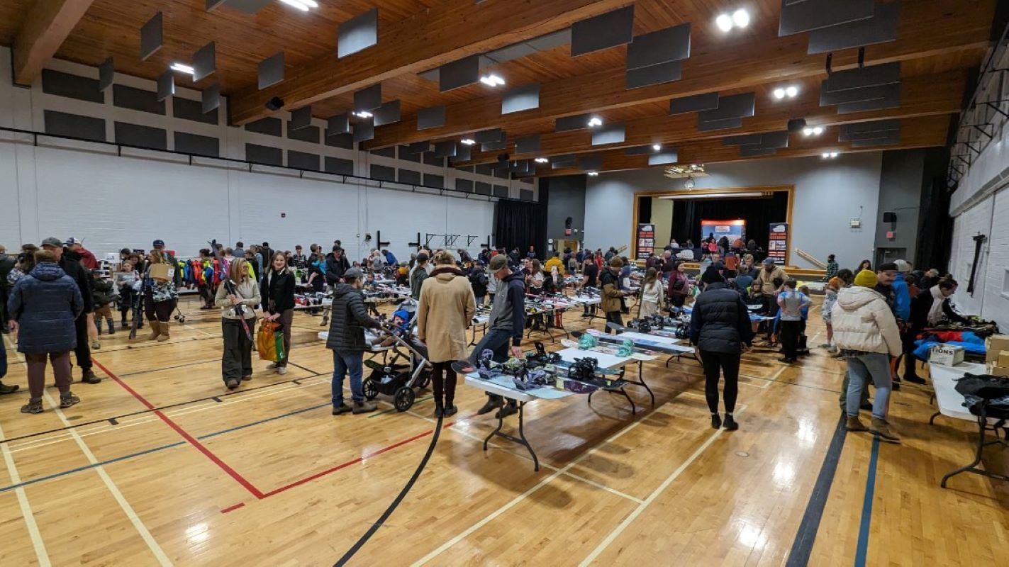 Crowded ski swap event in a gym with tables of winter gear and people browsing.