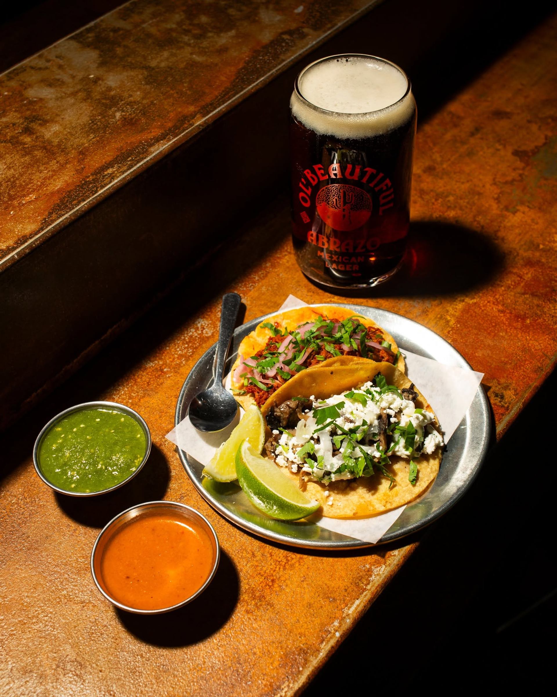 Tacos served with lime wedges, green and red salsa, sit beside a glass of craft beer.
