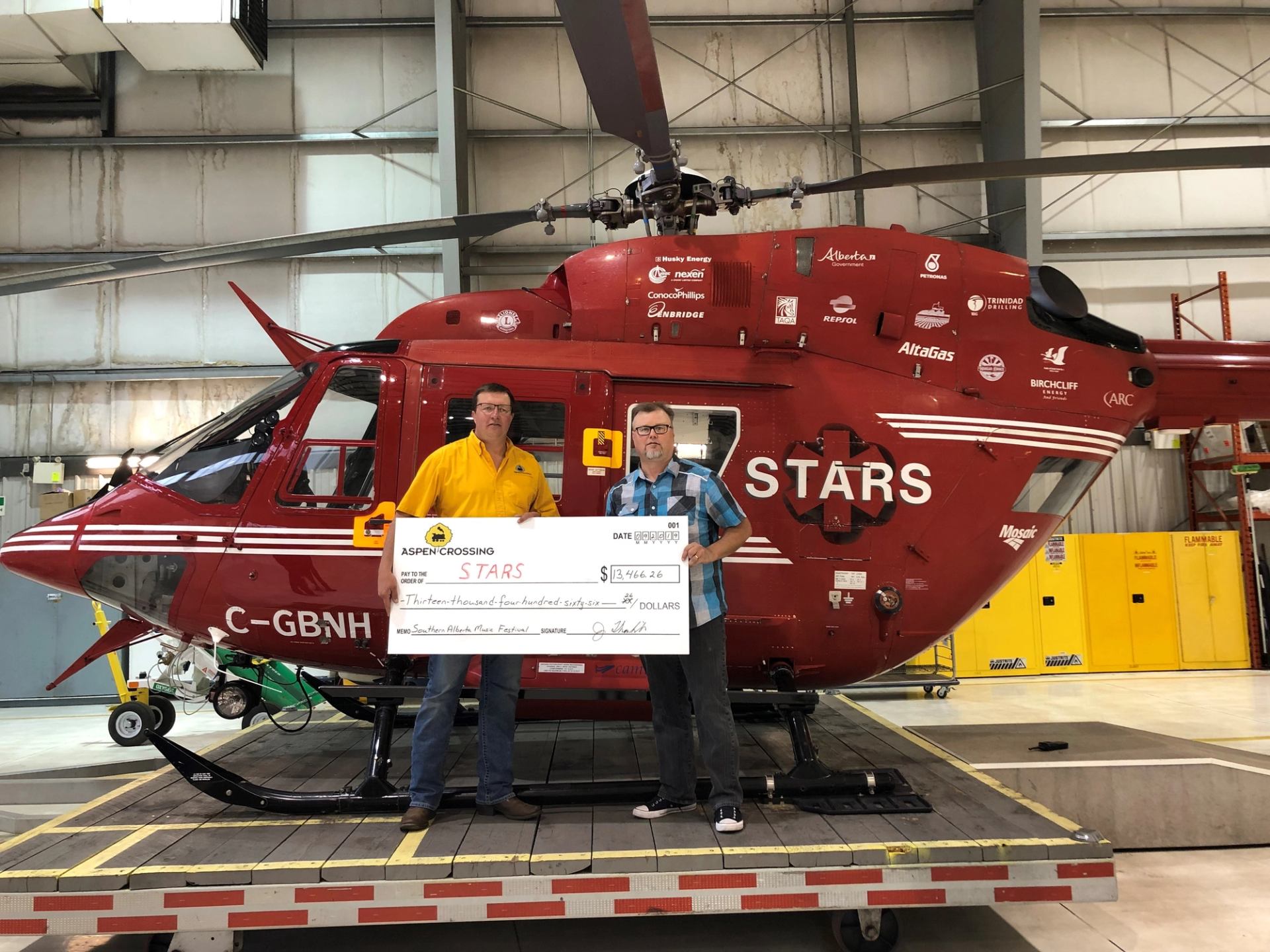 Red helicopter inside hangar; two men hold a cheque in front of cockpit window.