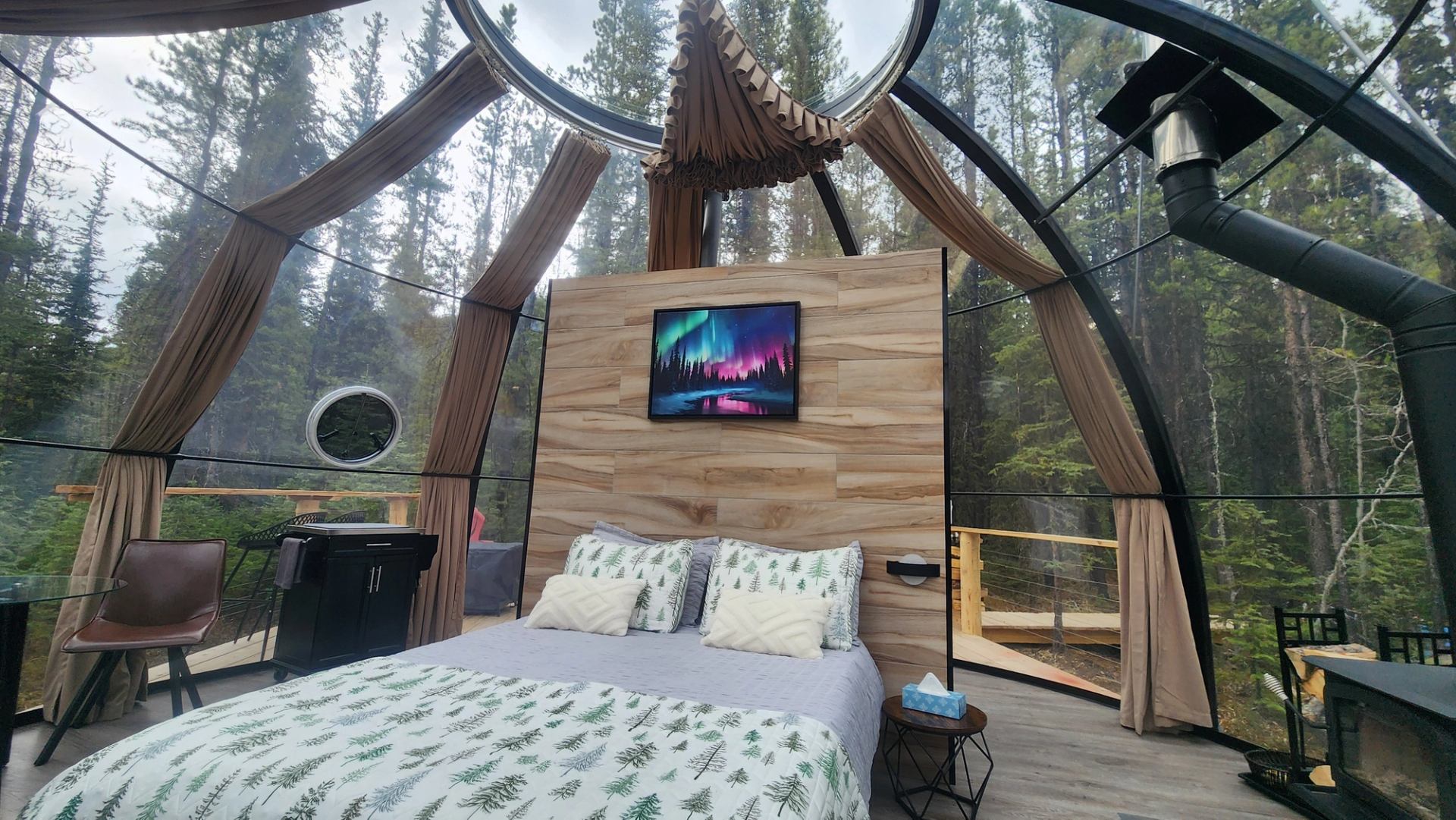 Glass-domed forest cabin bedroom with patterned bedding, wood headboard and aurora artwork.