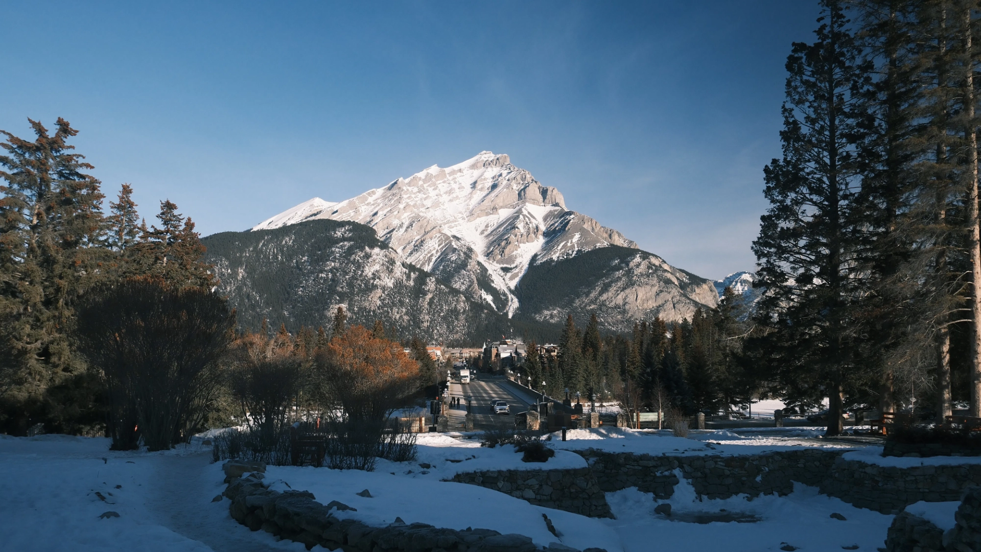 Snow-covered mountain rising behind forested valley and road with vehicles and buildings.