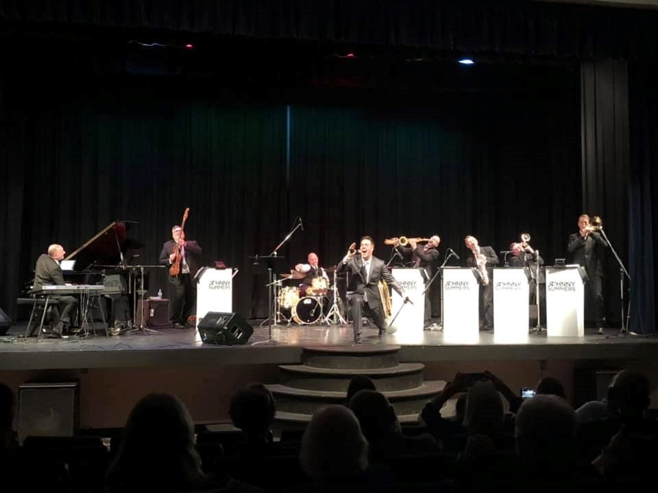 Stage performance with seated musicians, trumpets and saxophones, and microphones behind white riser.