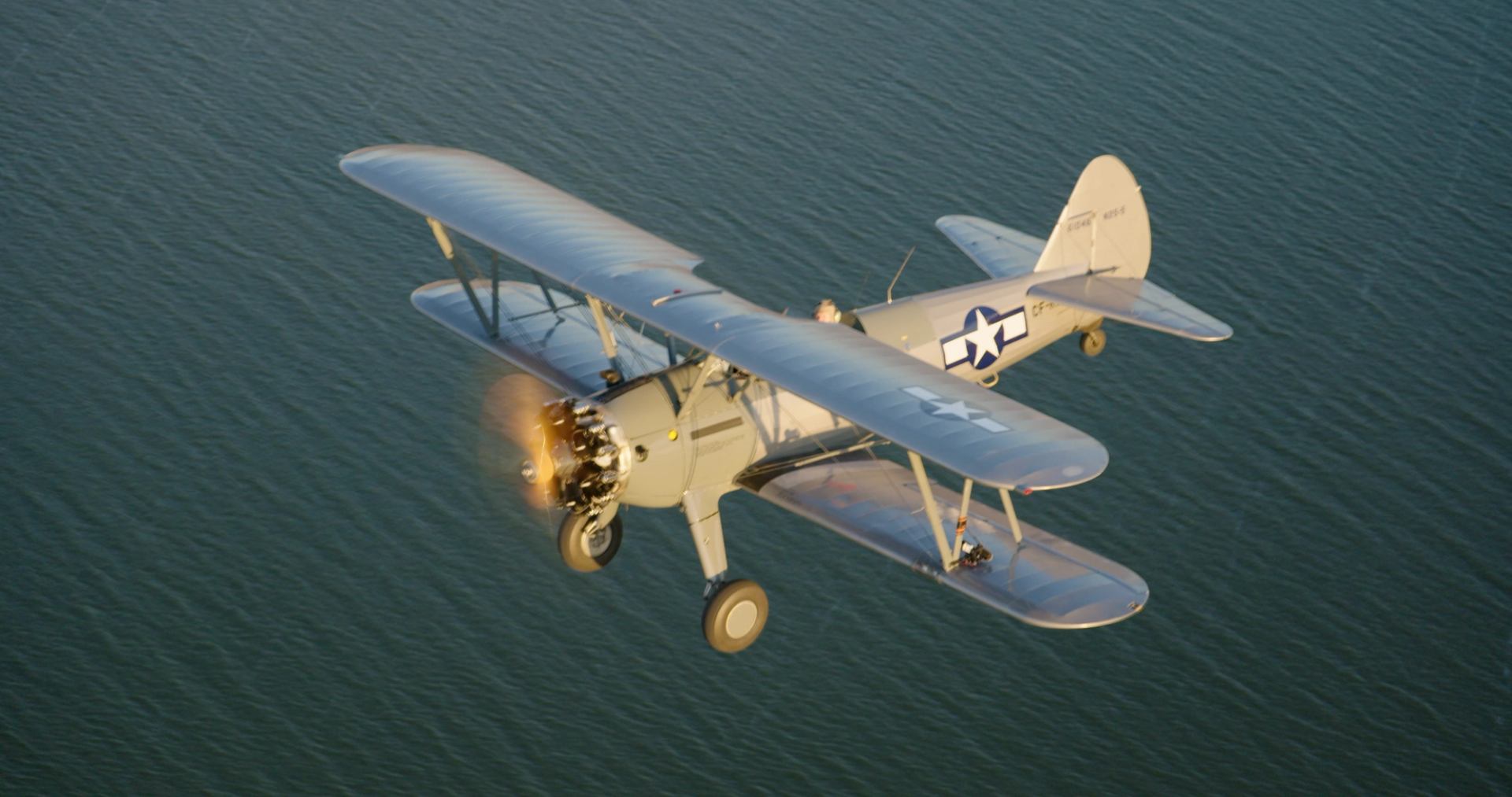 Vintage biplane with spinning propeller flies over calm water, showing military markings.