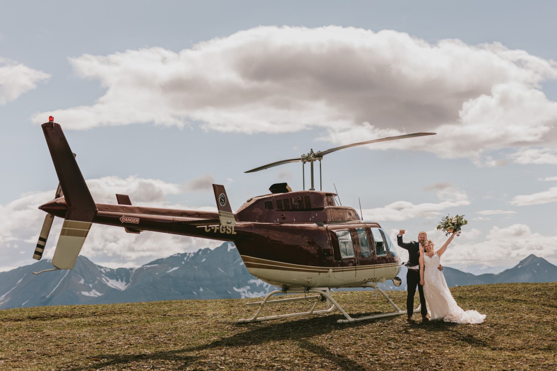 A bride and groom celebrate beside a helicopter on an open alpine plateau.