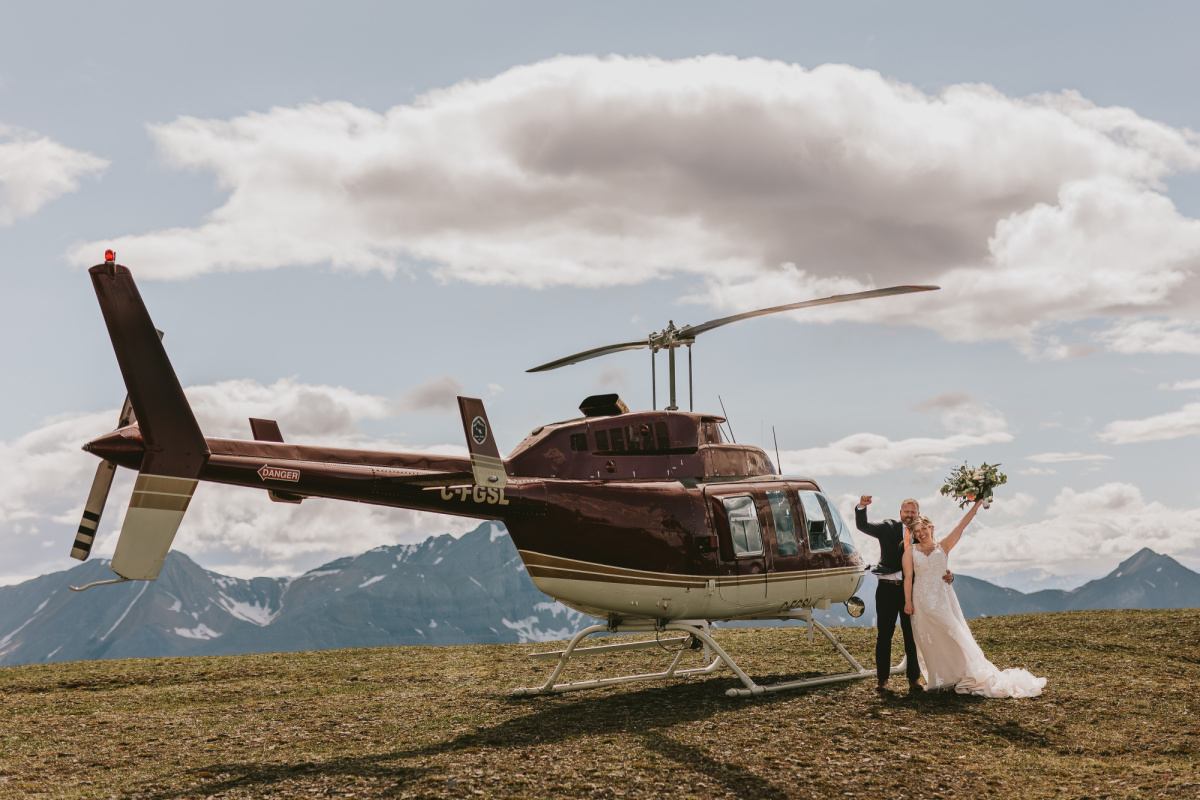 A bride and groom celebrate beside a helicopter on an open alpine plateau.