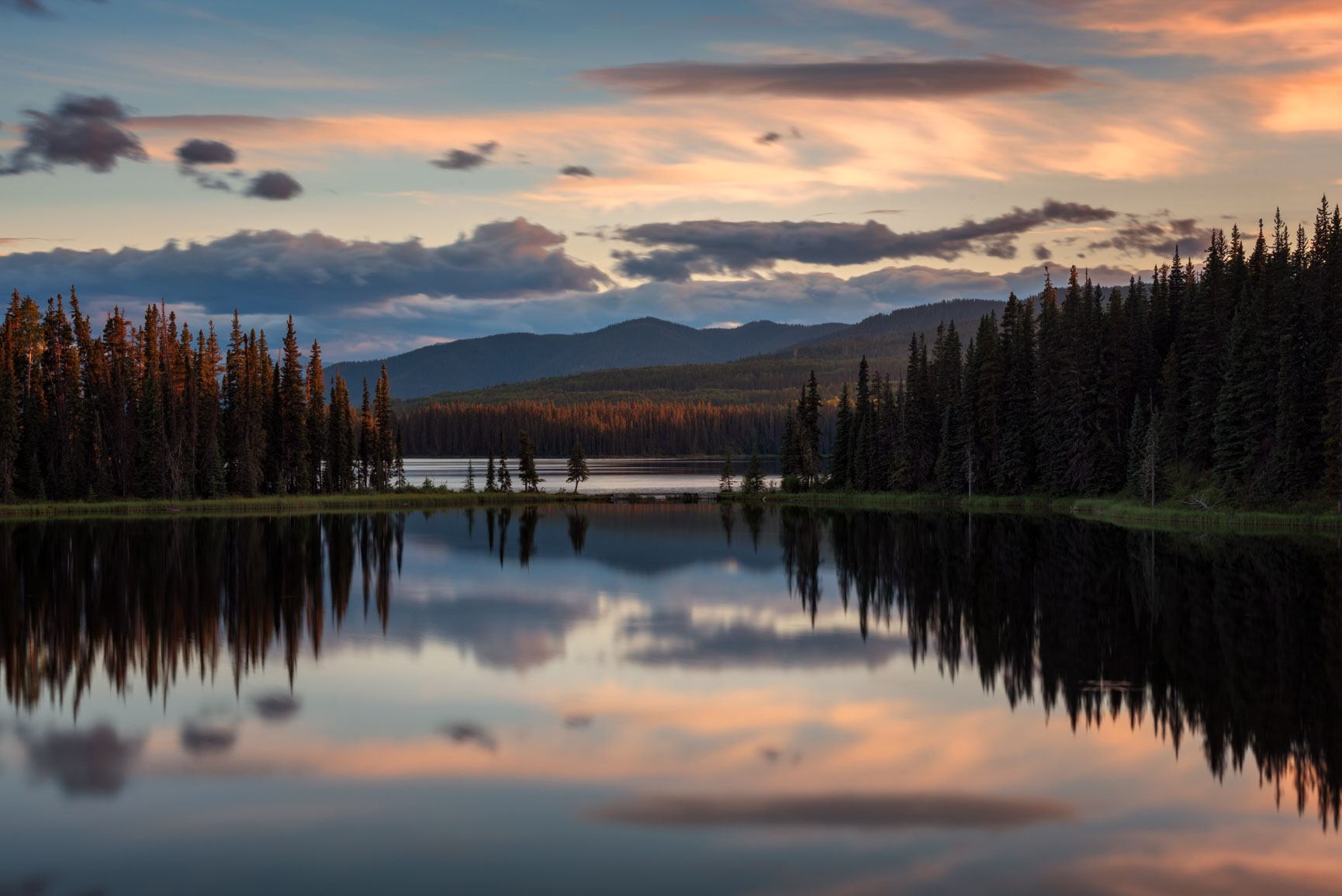Conifer forest and distant hills reflected in calm lake under colourful clouded sky.