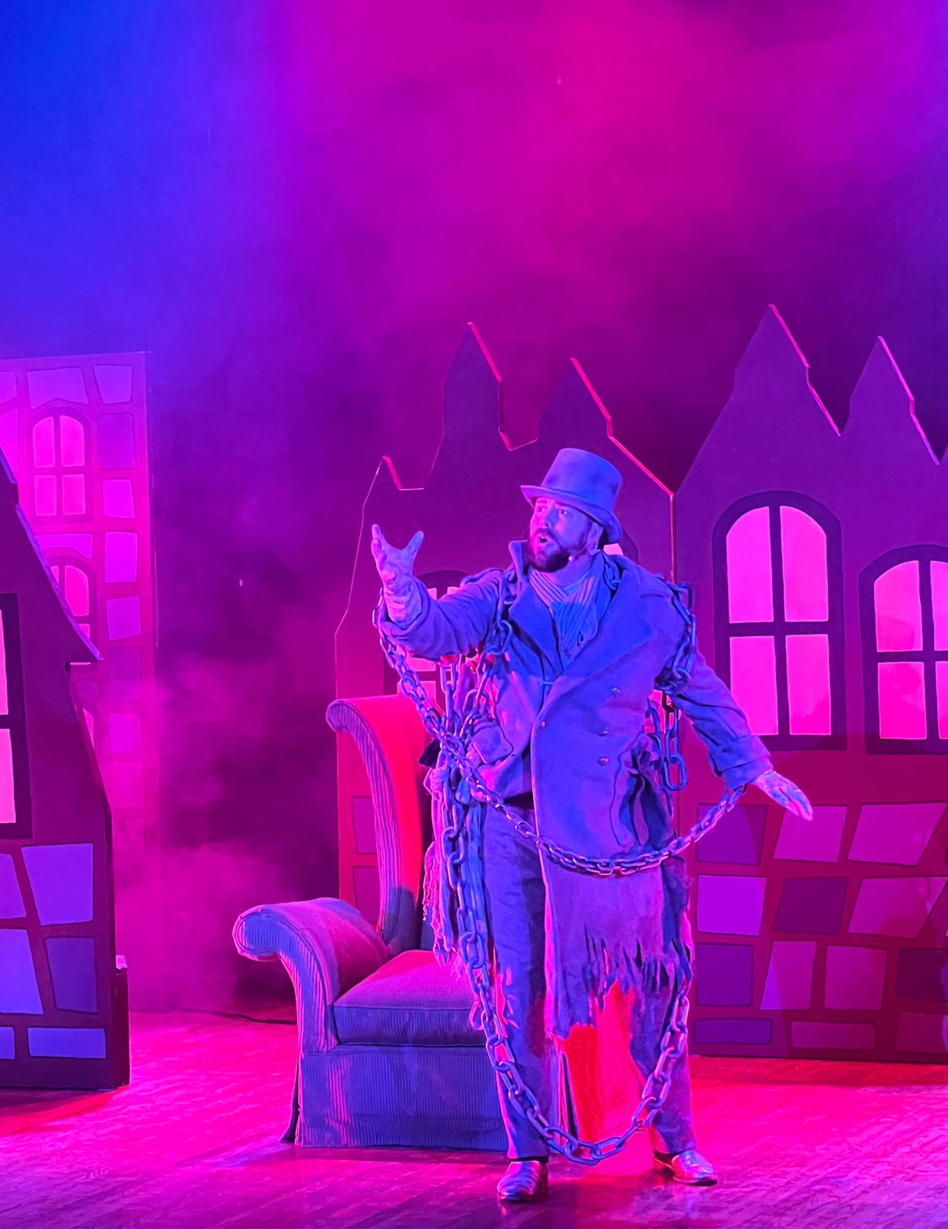 Actor in chains performs on stage with vibrant, colorful village backdrop.