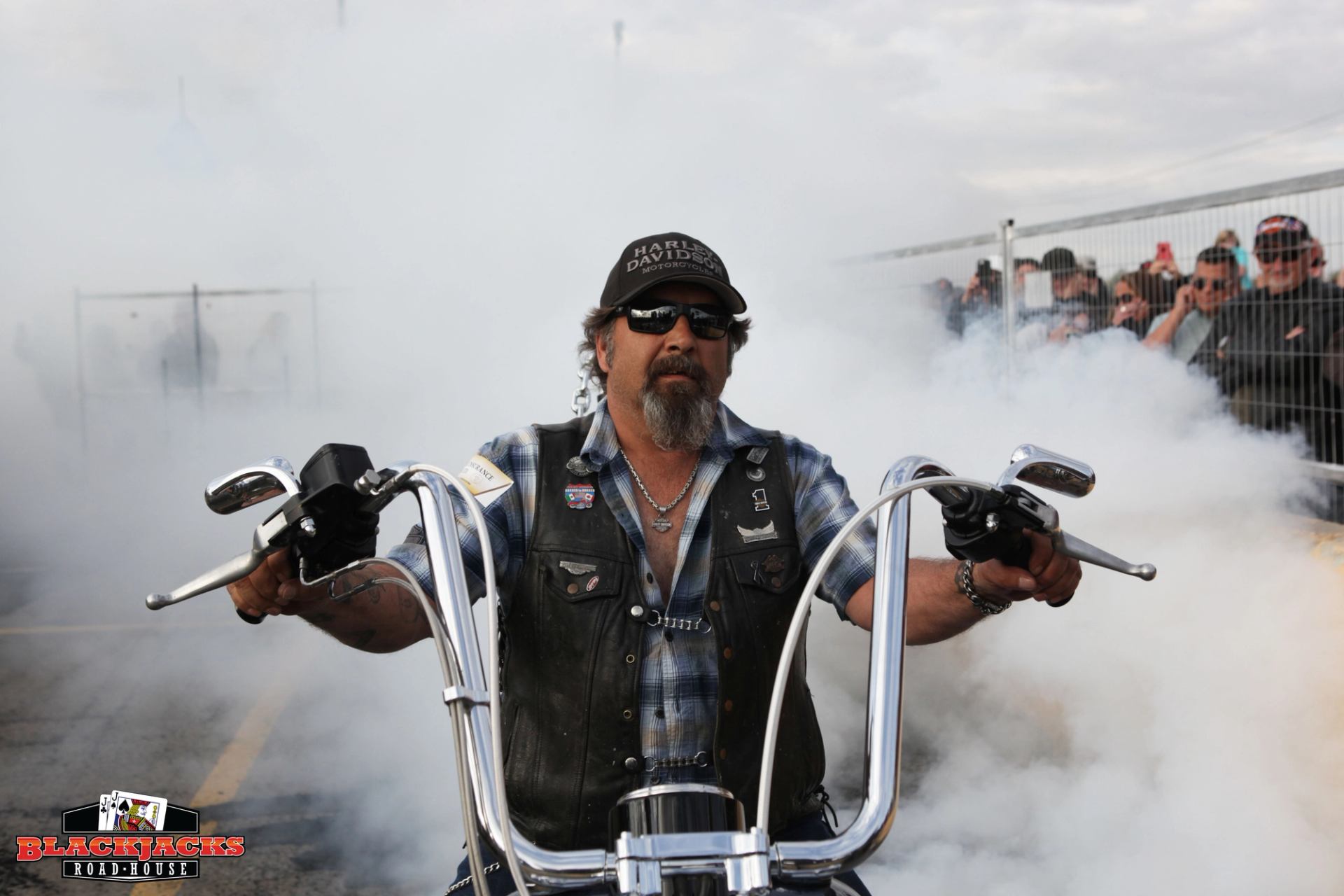 Motorcyclist in black vest and cap, holding chrome handlebars amid smoke at track event.