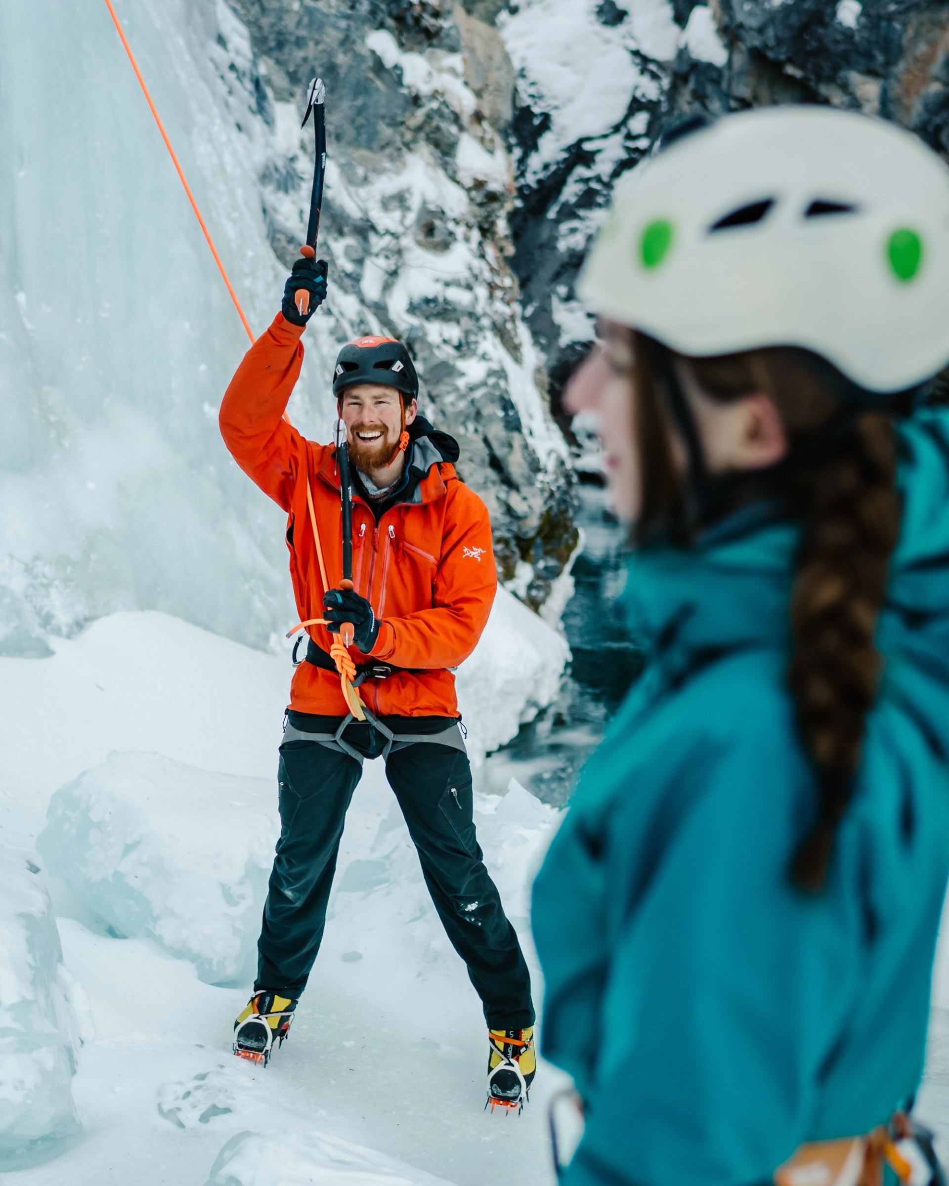 A smiling ice climber in an orange jacket holds an ice axe aloft, looking towards another climber in a snowy landscape.