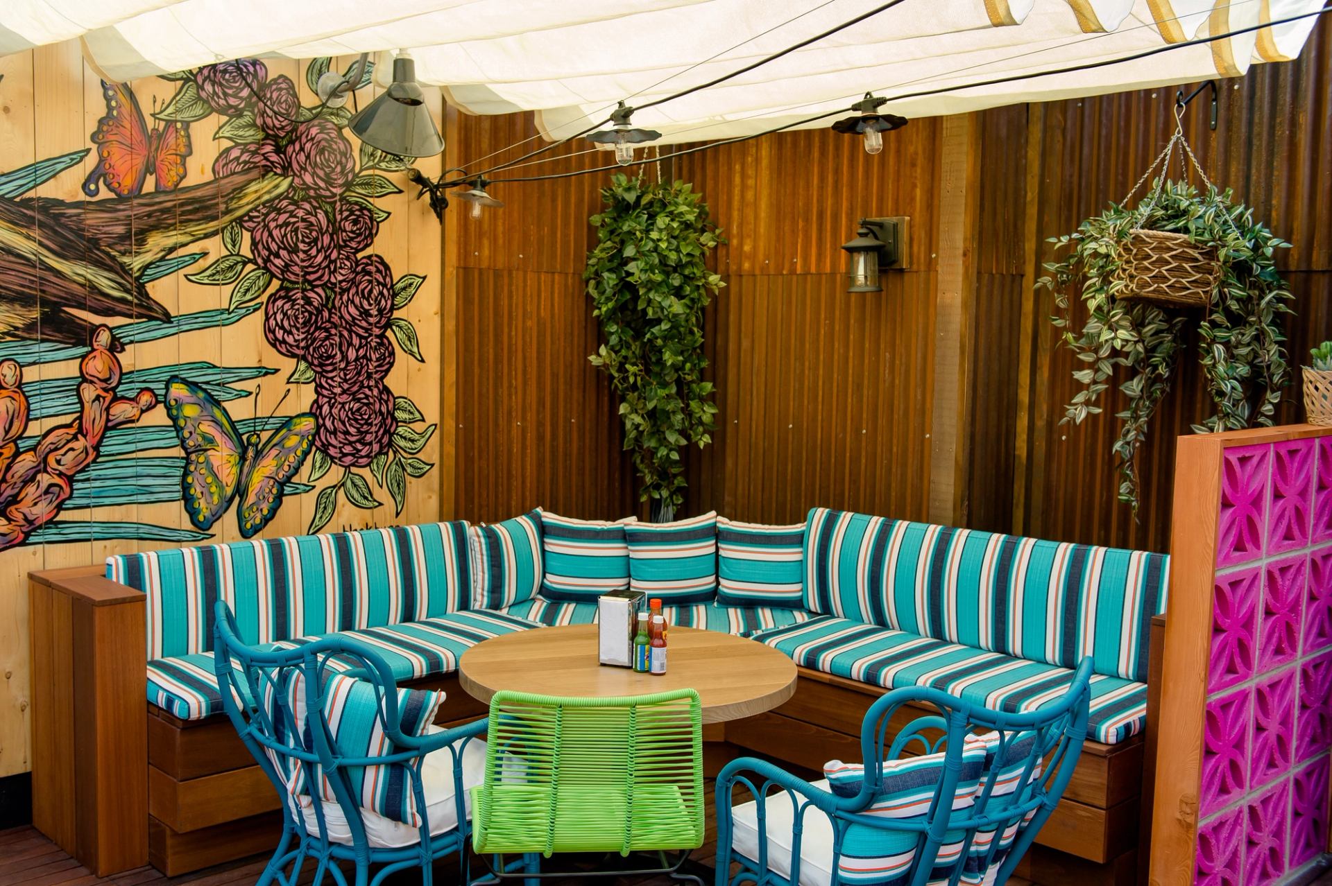 Vibrant outdoor patio with an L-shaped bench featuring turquoise striped cushions, a round table, a butterfly and flower wall mural, and hanging plants.