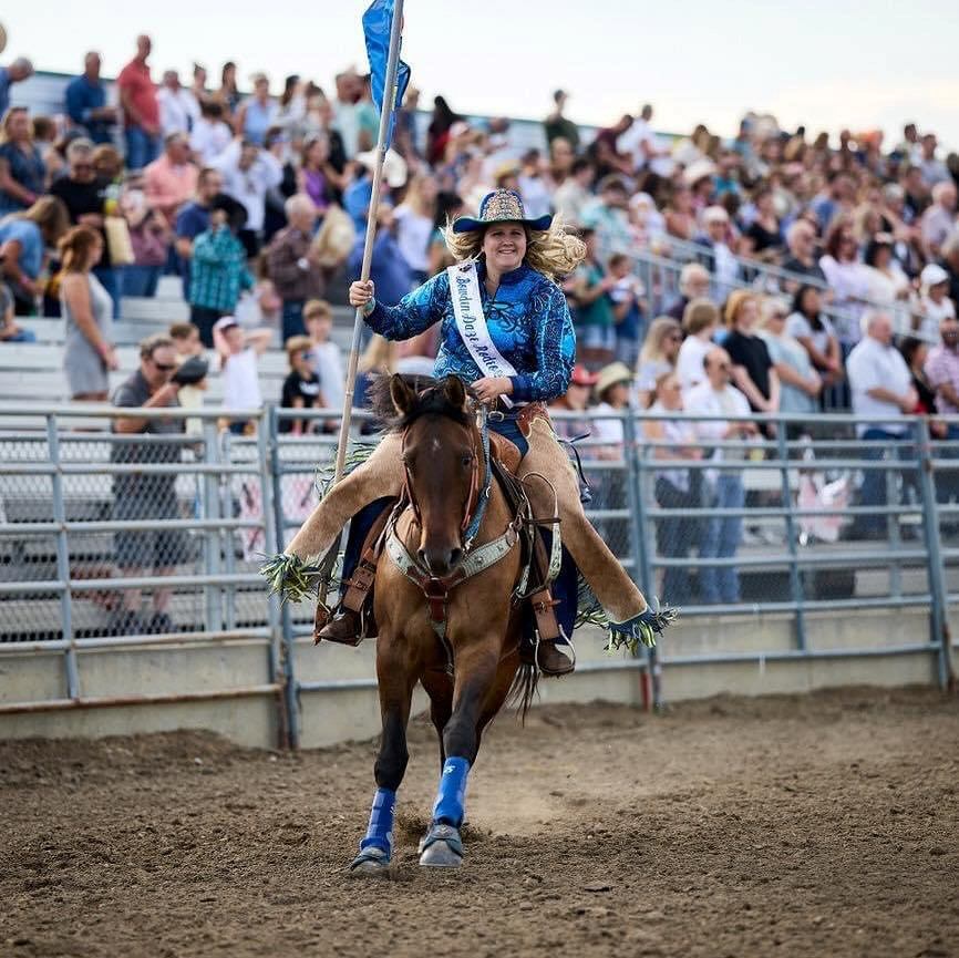Rodeo queen rides a horse in a rodeo arena holding a blue flag.